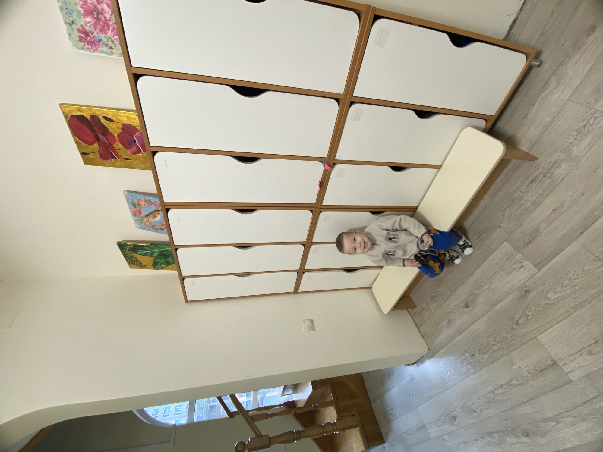 A low-angle indoor shot features a young boy seated on a white surface, adjacent to a tall, rectangular storage unit with multiple white drawers framed in brown. The storage unit has colorful floral and botanical-themed decorative panels attached to the side. The boy is wearing a light-colored sweatshirt and blue pants. The floor is light gray and wood-patterned. A portion of a wooden staircase is visible in the background. The location is in Sochi, Russia.