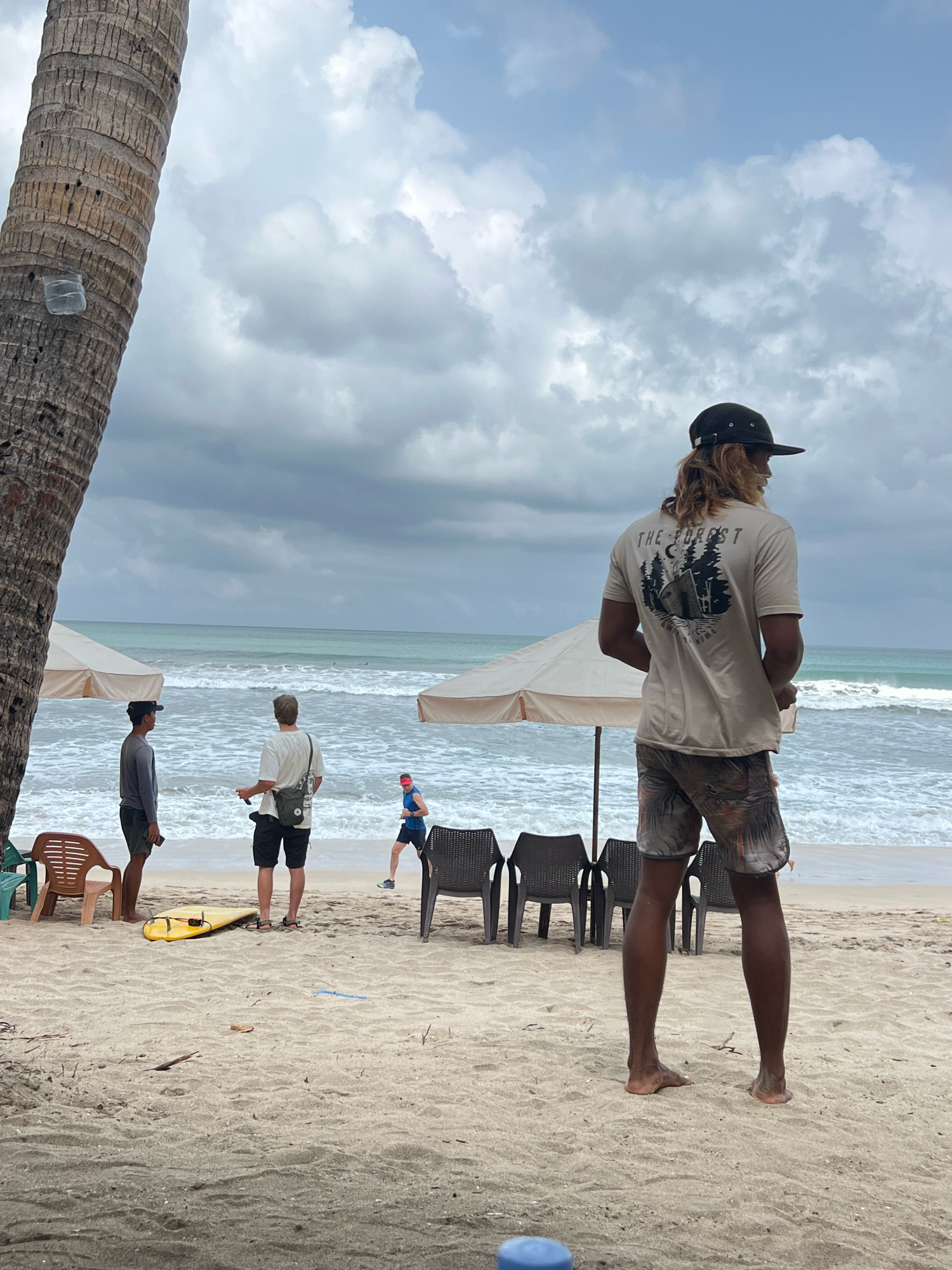 The image depicts a beach scene in Kuta, Indonesia, under a cloudy sky. In the foreground, a person wearing a dark baseball cap and a light-colored t-shirt with a graphic print of a tent and trees, along with patterned shorts, stands facing away from the camera, looking towards the ocean. Behind them, two other individuals are visible on the sand. One, wearing a grey long-sleeved shirt and dark shorts, stands to the left. Another, in a white t-shirt and black shorts, stands slightly to the right of the first person. Further back, a person in a blue tank top and dark shorts with a red headband is running on the beach. Several empty beach chairs and two large umbrella-like structures are set up on the sand. A yellow surfboard lies on the ground near the chairs. The ocean, with visible waves, stretches out to the horizon. A coconut palm tree trunk is prominent on the left side of the image.
