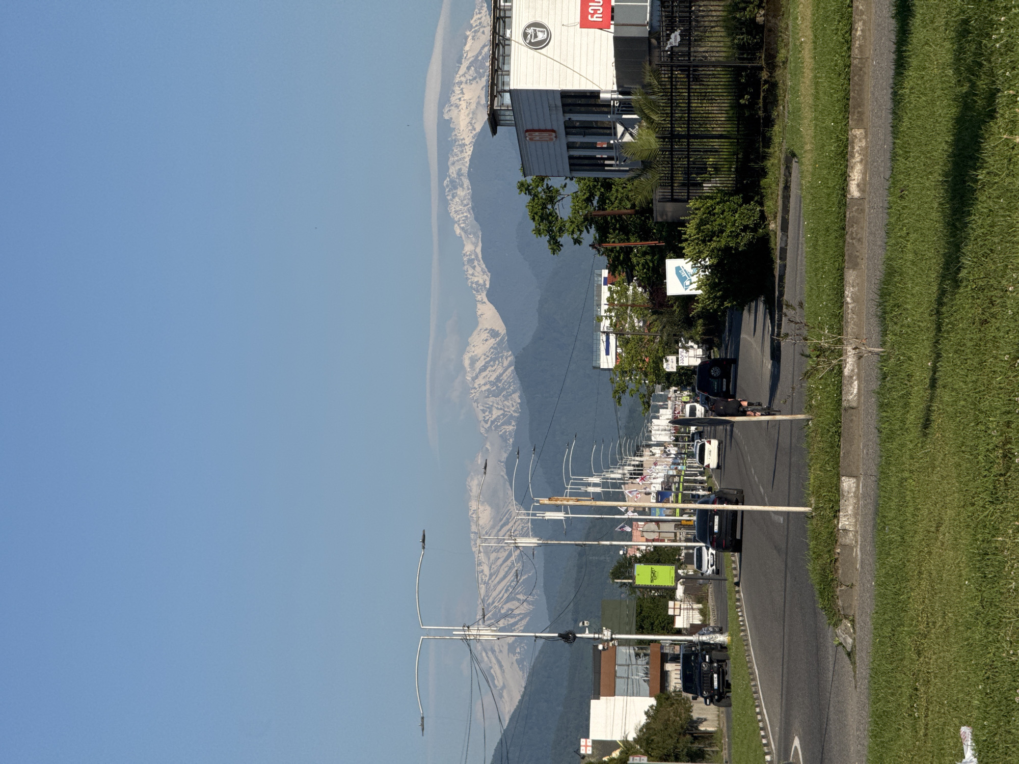 The image shows a street scene in Batumi, Georgia. The street runs perpendicular to the horizon, with a grassy area and sidewalk on the right and buildings on the left. Cars are visible on the street. A row of streetlights lines the street. In the background, mountains are visible against a clear blue sky. Buildings and various signage are also present along the street.
