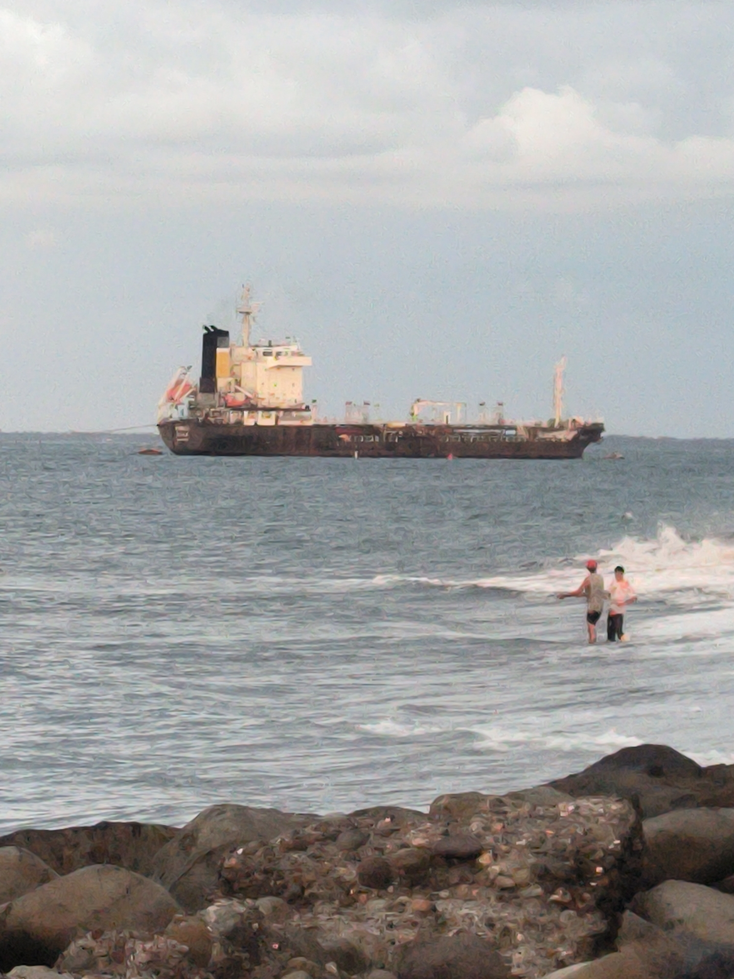 The image depicts a wide shot of the sea in Lhokseumawe, Indonesia. In the background, a large cargo ship is anchored offshore. In the middle ground, two individuals are wading in the shallow water near the shore, which is lined with rocks. The sky is overcast, contributing to a muted color palette. The overall impression is one of a calm, perhaps slightly dreary, day by the sea.