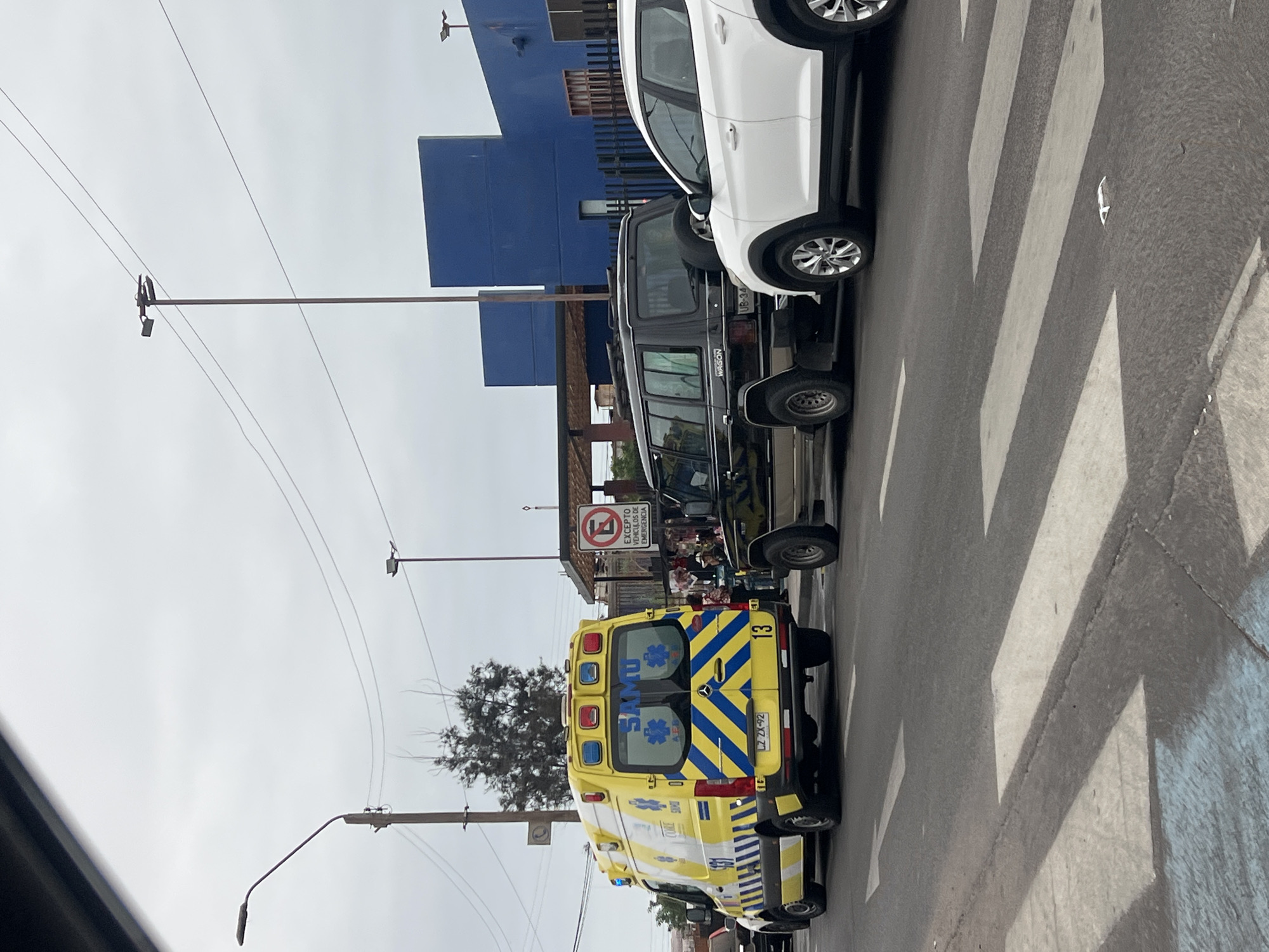 The image presents a street scene in Arica, Chile, under an overcast sky, likely during the daytime. The perspective is from inside a vehicle, looking towards the rear of a yellow and blue ambulance stopped on the right side of the road.
The ambulance, belonging to the SAMU (Servicio de Atención Médica de Urgencia) service, features "SAMU" emblazoned on its rear doors, along with blue and yellow striped markings. It has an emergency light bar on its roof and the number "13" on its right rear. Its Chilean license plate, "LZ ZX-92," is visible.
Further down the street, in front of the ambulance, are two other stationary vehicles. A black utility vehicle or SUV, marked with "WAGON" on its rear, displays the license plate "UB-34." Beyond it, a white SUV is also visible.
The asphalt road features a distinct pedestrian crosswalk with white stripes. On the right side of the street, a building with a vibrant blue facade is partially seen. A red circular street sign indicates "No Stopping/Parking" (a red circle with a diagonal line), with the text "EXCEPTO VEHÍCULOS DE EMERGENCIA" (EXCEPT EMERGENCY VEHICLES) written below, justifying the ambulance's position. Overhead, power lines and a streetlight pole are visible against the cloudy sky. A small tree with sparse foliage stands in the mid-ground. The scene appears static, with no discernible active event or people.