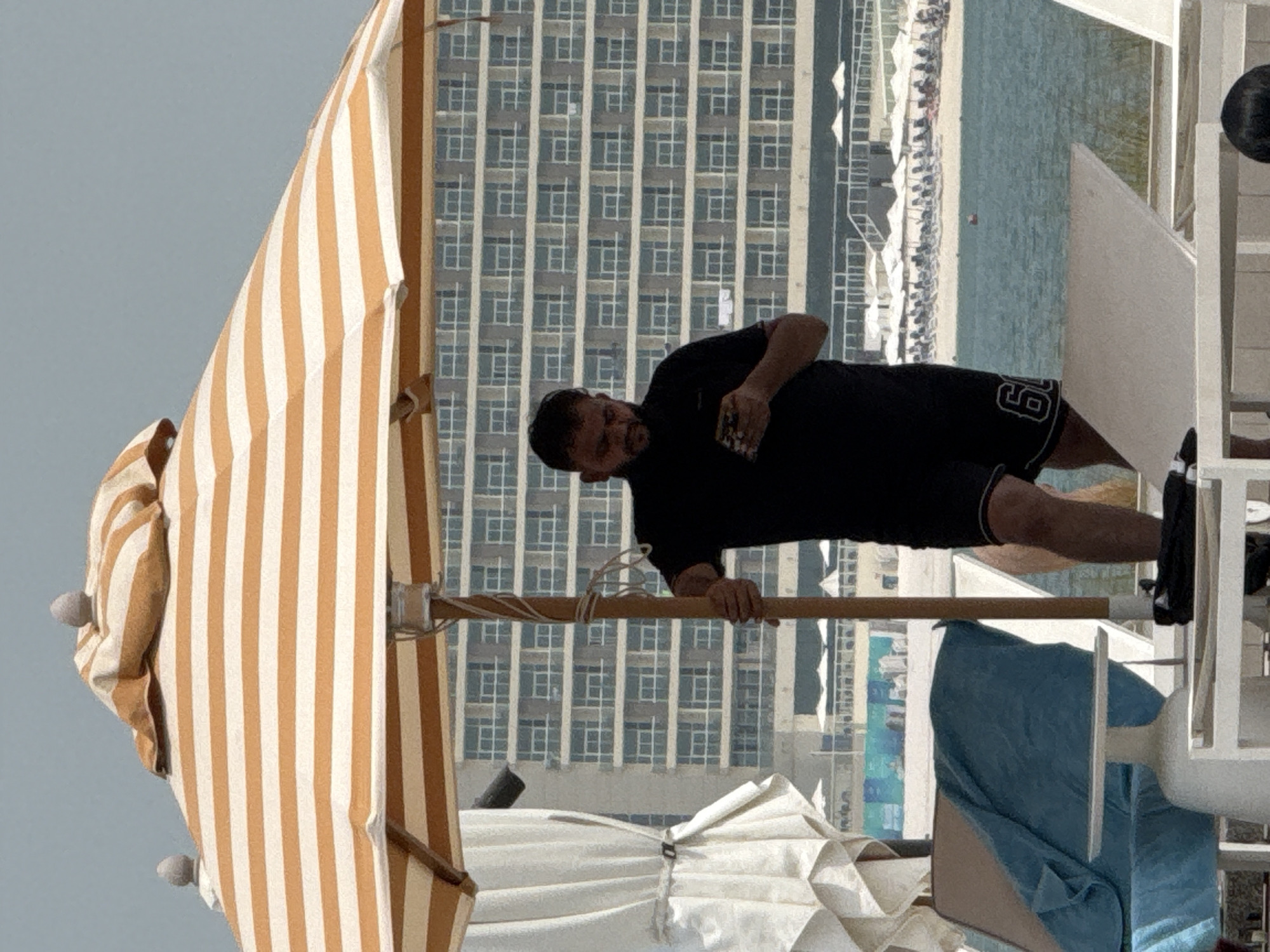 A man wearing a black t-shirt and black shorts with white trim is standing under an orange and white striped umbrella on a beach. He is holding a cell phone in his right hand and looking down at it. The man has dark hair and a beard. In the background, there is a large, modern hotel building with many windows. There are also beach chairs and umbrellas set up on the beach. The water is a light blue color. The scene appears to be in the city of Ras Al-Khaimah, United Arab Emirates.