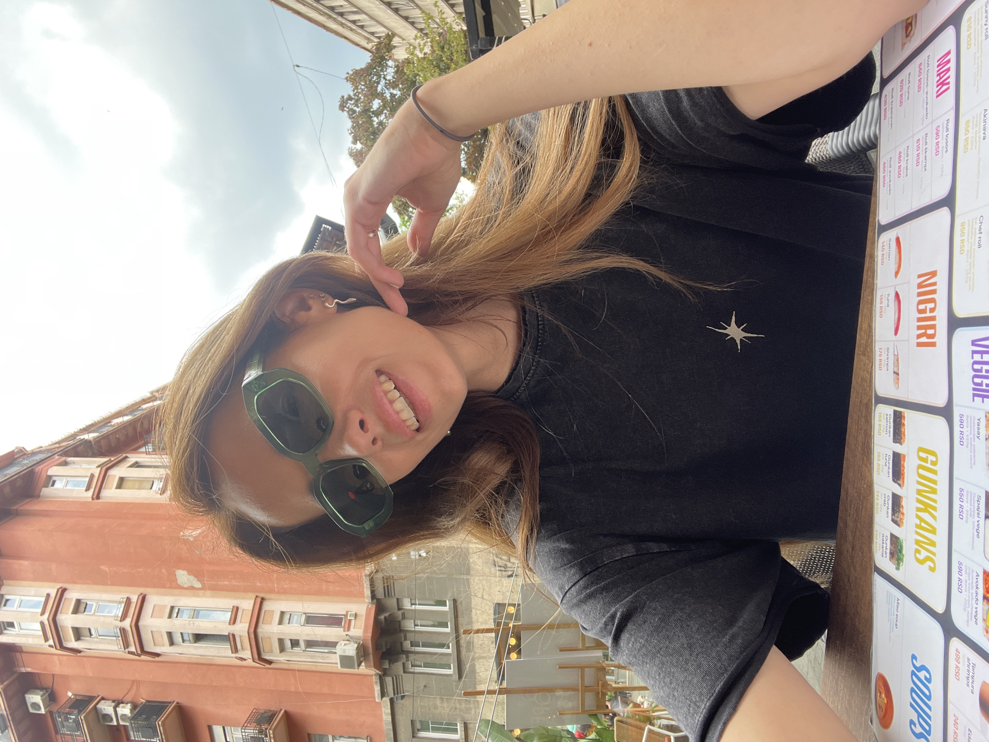 The image shows a woman taking a selfie at an outdoor restaurant in Belgrade, Serbia. The subject is fair-skinned and is wearing sunglasses and a black t-shirt with a white star emblem. She is smiling and has long hair, accessorized with a snake-shaped earring. The background reveals typical Belgrade architecture and a menu featuring sushi and other Asian cuisine. The image captures a casual, urban setting.