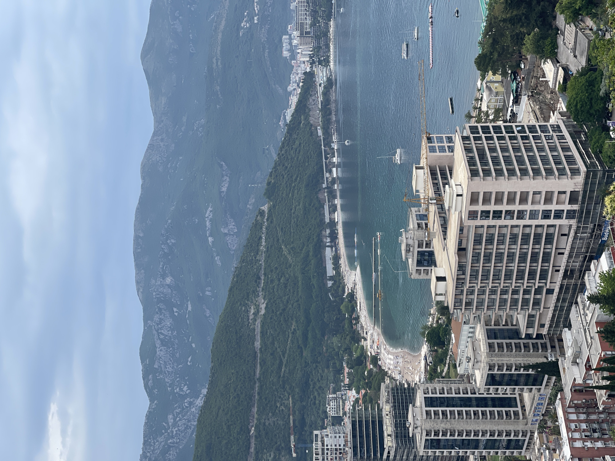 The image depicts a view of the city of Budva, Montenegro, featuring a coastline with beaches and buildings. Visible are mountains in the background, a sea with boats, and modern architectural structures in the foreground. There are people populating the beach areas, umbrellas are visible, and some construction is taking place involving cranes next to buildings.
