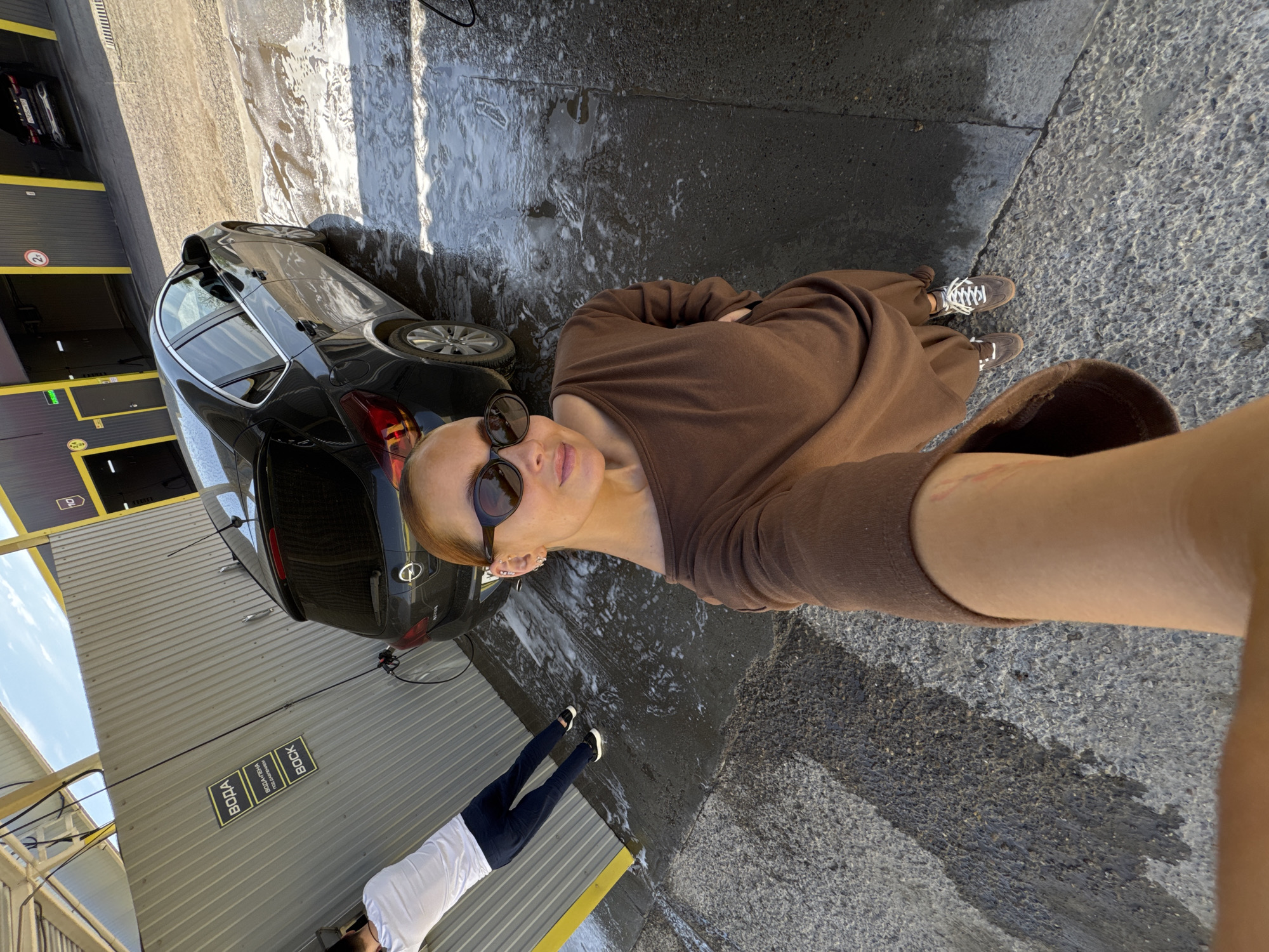 The image shows a person in a brown dress and sunglasses taking a selfie at a car wash in Chaikovsky, Russia. A black car is partially visible on the left, positioned near the entrance of one of the car wash bays. Another person wearing a white shirt and dark pants is walking in the background near a gray corrugated metal wall with Cyrillic text. The ground is a mix of wet and dry pavement.