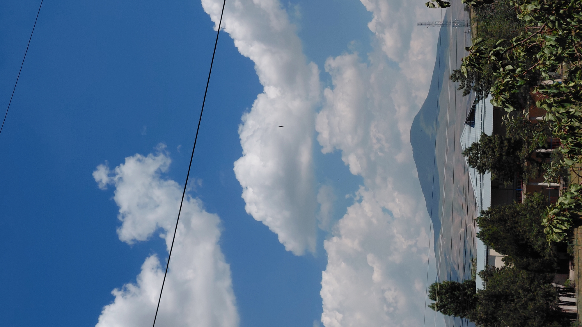 The image, taken from a distance, depicts a rural landscape under a bright blue sky with scattered white clouds. In the foreground, lush green trees and foliage are visible. Behind the trees, a building with a light blue roof and light-colored walls can be seen. Further in the background, a large, hazy mountain dominates the horizon. A power line stretches across the frame, bisecting the sky. The scene appears to be located in or near Abovyan, Armenia, suggested by the characteristic mountainous terrain and architecture. There are no people or animals clearly visible in the frame. The overall impression is one of a tranquil, natural setting.