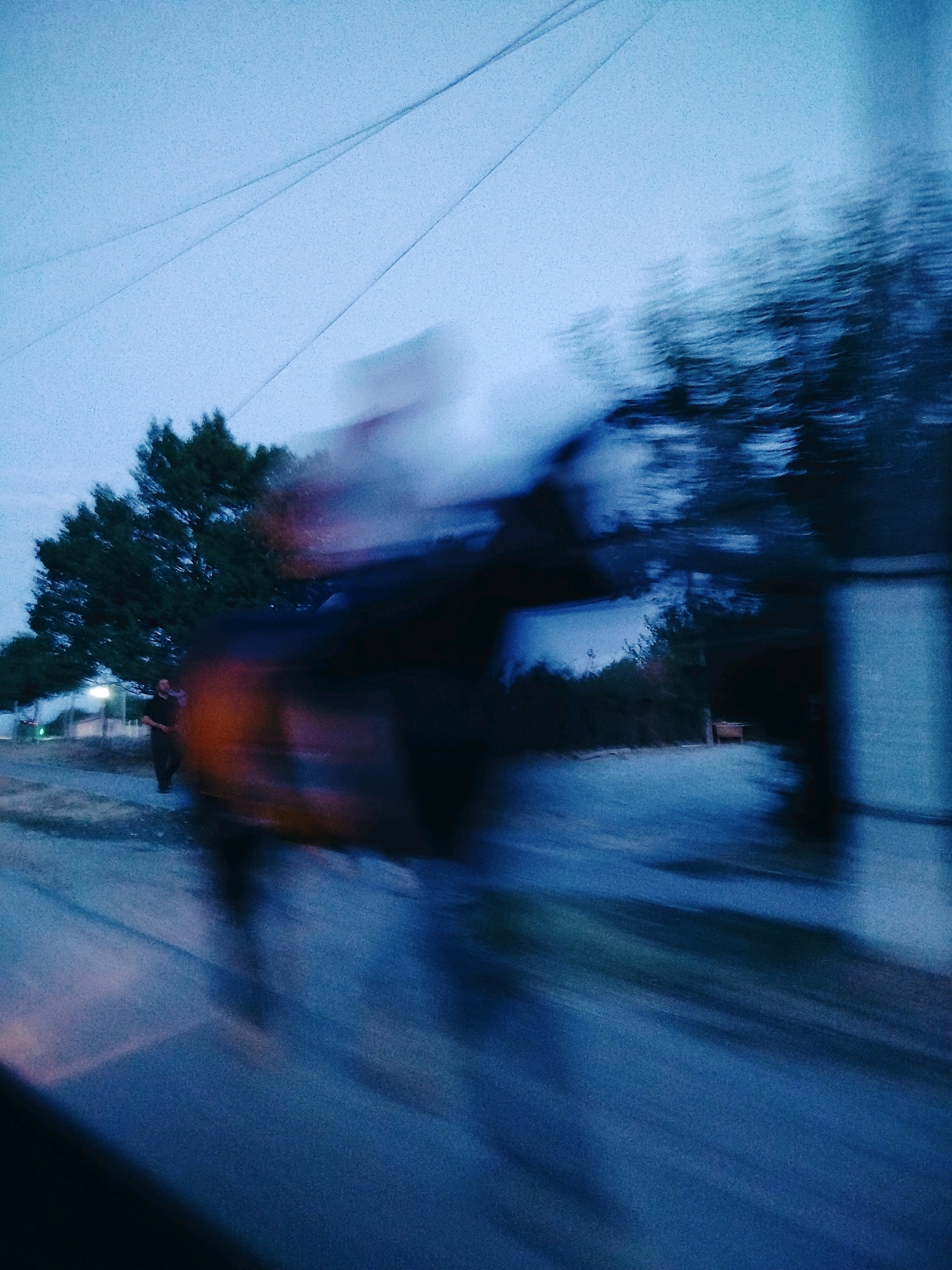 The image captures a blurry, motion-filled scene at dusk or dawn, possibly on a street or road. A lone figure, appearing to be a man in dark clothing, stands on the side of the road near a tall tree. The foreground is dominated by streaks of blue and dark colors, suggesting movement, possibly from a vehicle or a fast-paced camera. The background shows the silhouette of trees and a building or structure. The sky is a pale, hazy blue. The overall impression is one of a fleeting moment, captured with intentional blur, evoking a sense of movement and perhaps a feeling of isolation or transient observation. The location is identified as Kolkent, Kazakhstan.