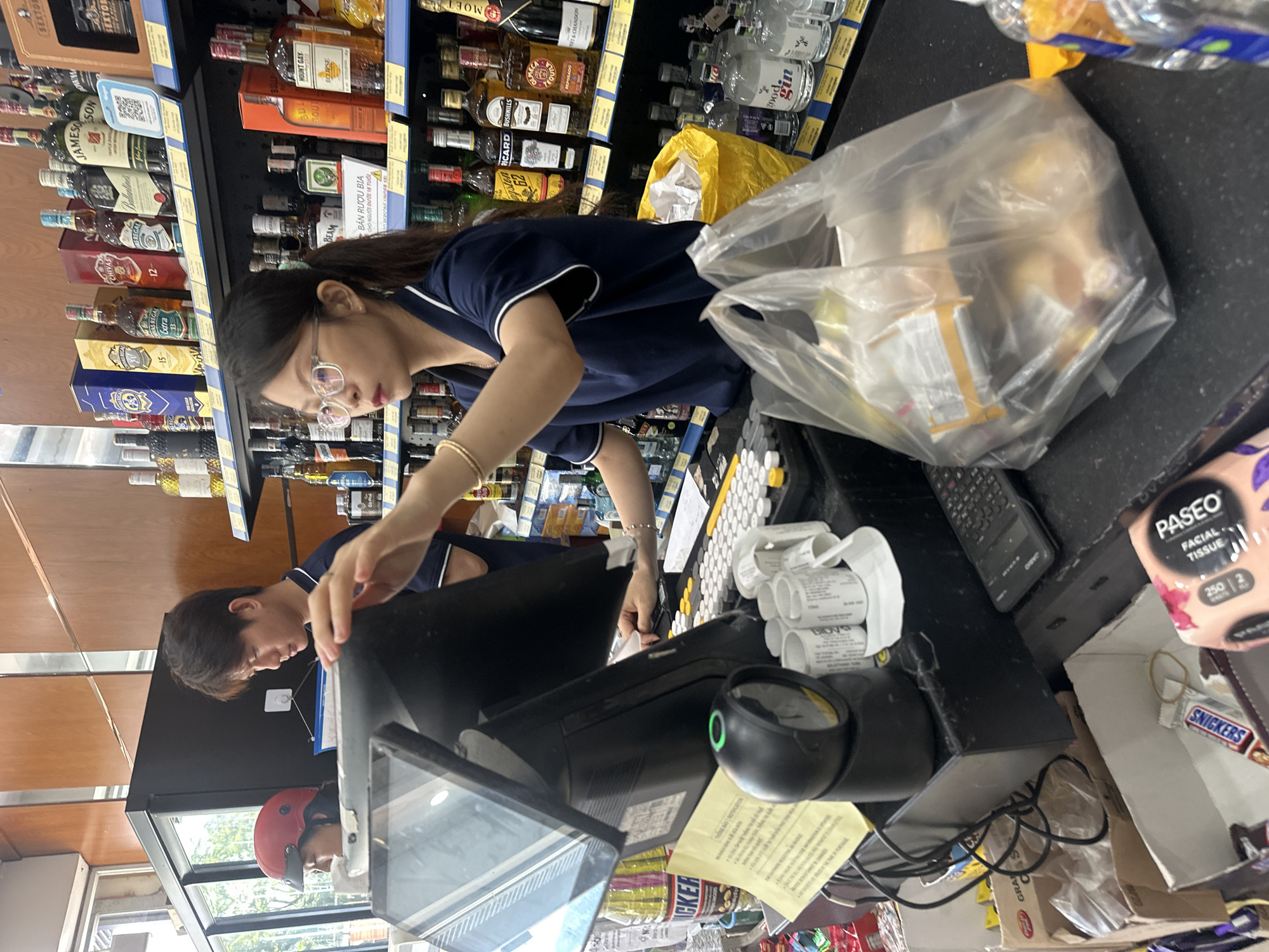 In Bac Nha Trang Ward, Vietnam, two individuals are visible at a checkout counter in a retail establishment. To the left, a person wearing a red helmet and dark clothing is partially visible, facing towards the right. Adjacent to this person, a young man with dark hair, wearing a dark blue collared shirt with white trim, stands behind the counter. He appears to be looking down and to the right.
In the foreground, a young woman with glasses and dark hair pulled back is operating a touchscreen point-of-sale system. She is wearing a dark blue collared shirt with white trim and a gold bracelet on her left wrist. Her right hand is extended towards the screen, and her left hand is resting on the counter. In front of her, a black scanner with a green light is positioned on the counter. Various receipts are scattered around the scanner, along with a keyboard and several small white containers. A clear plastic bag filled with items is placed on the counter in the foreground, obscuring a yellow bin. Behind the woman, shelves are stocked with numerous bottles of alcohol. A calculator is situated to the right of the keyboard. The overall environment suggests a convenience store or a similar retail setting.