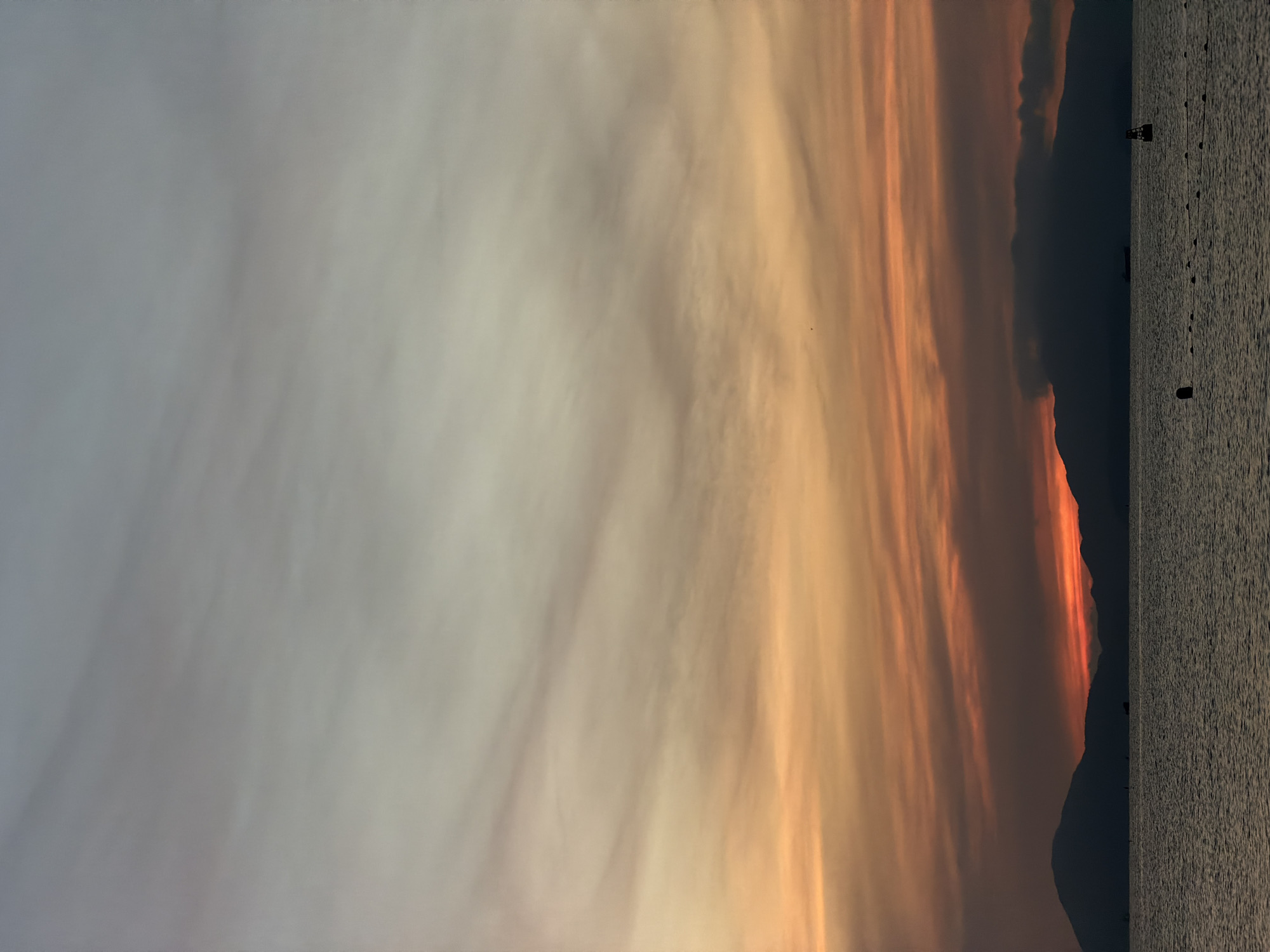 The image shows a sunset over the sea near Hoi An, Vietnam. The sky is filled with hues of orange, pink, and pale blue, with wispy clouds streaked across it. Silhouetted mountains form the horizon line. The sea is calm, with gentle ripples on the surface. In the distance, several boats are visible, including a large cargo ship and a smaller buoy. The scene is serene and peaceful, conveying a sense of natural beauty and tranquility.