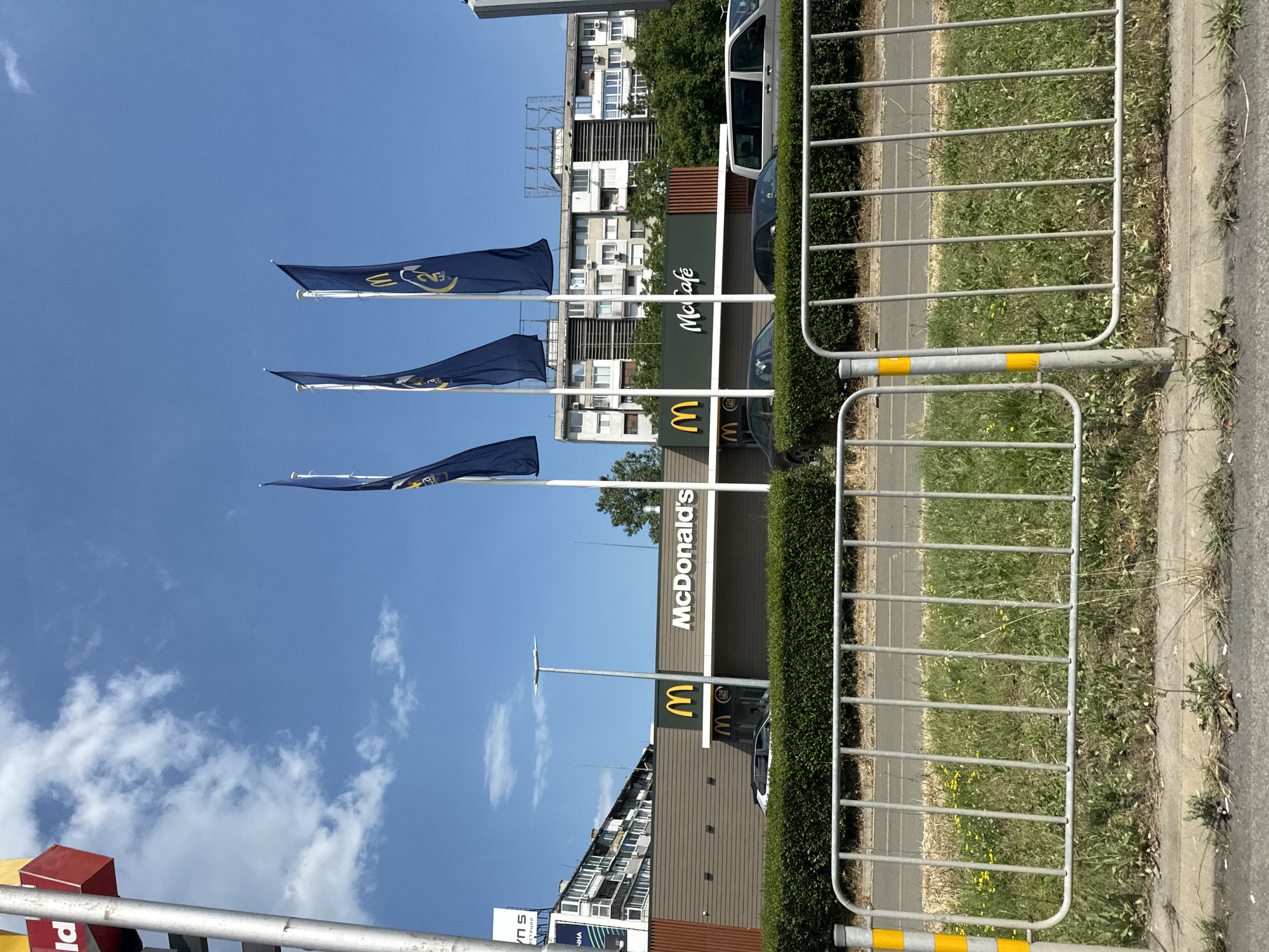 The image shows a McDonald's restaurant in Burgas, Bulgaria, on a sunny day with a blue sky and scattered clouds. The restaurant is a modern building with a brown facade and a large McDonald's logo. Three blue flags with yellow McDonald's branding are flying in front of the restaurant. A McCafé sign is also visible. Several cars are parked in front of the restaurant, and a hedge runs along the edge of the property. In the background, there are residential buildings and trees. A metal fence with yellow and white stripes is in the foreground, separating the street from the restaurant. The image appears to have been taken from across the street.