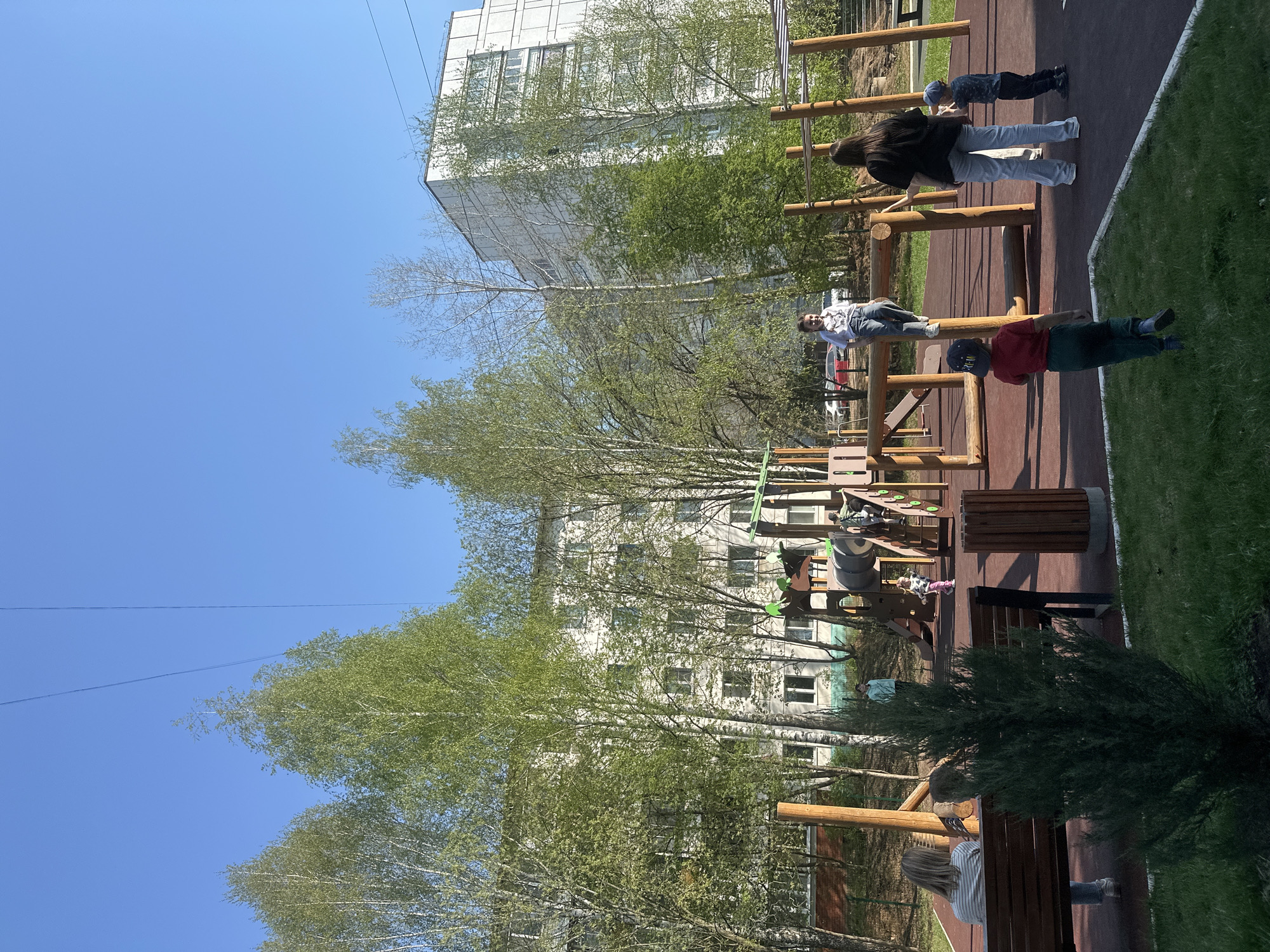 The image shows an outdoor playground in Chaikovsky, Russia, with a series of wooden exercise structures. Several people are using the equipment, including adults and children. A multi-story building is visible in the background, partially obscured by trees with green leaves. The sky is blue with no clouds. A bench is visible near the bottom right corner of the image, occupied by a person. The playground is a mix of reddish-brown surfaces and green grass. A power line is visible in the sky above the trees.