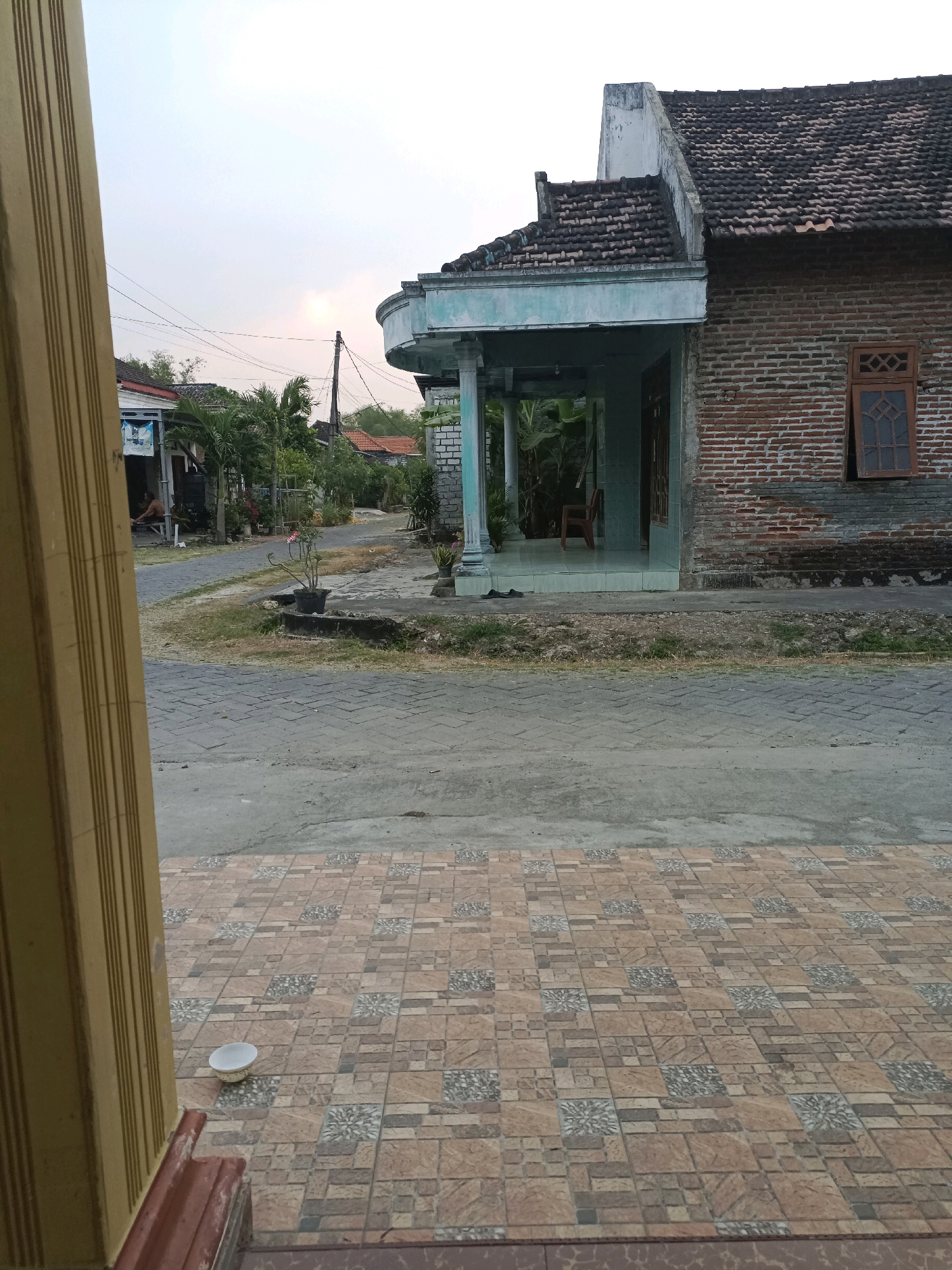 The image depicts a street scene in Rancang Kencono, Indonesia, likely during the early morning or late afternoon due to the soft lighting. In the foreground, a section of a building's facade is visible on the left, characterized by vertical wooden planks. The ground is paved with patterned square tiles, and a small white bowl sits on these tiles.
The street extends into the midground, where it curves slightly. A single-story house with a brick exterior and a dark tiled roof dominates the right side of the frame. The house features a covered porch with light blue trim and columns. Adjacent to the porch, a wooden chair is visible. A wooden-framed window with a distinctive geometric pattern is set into the brick wall.
On the left side of the street, several other houses are visible, some with palm trees and other greenery in front of them. Power lines stretch across the sky. A person is seated on a bench outside one of the houses on the left. The overall environment appears to be a residential area with a slightly worn or aged aesthetic.