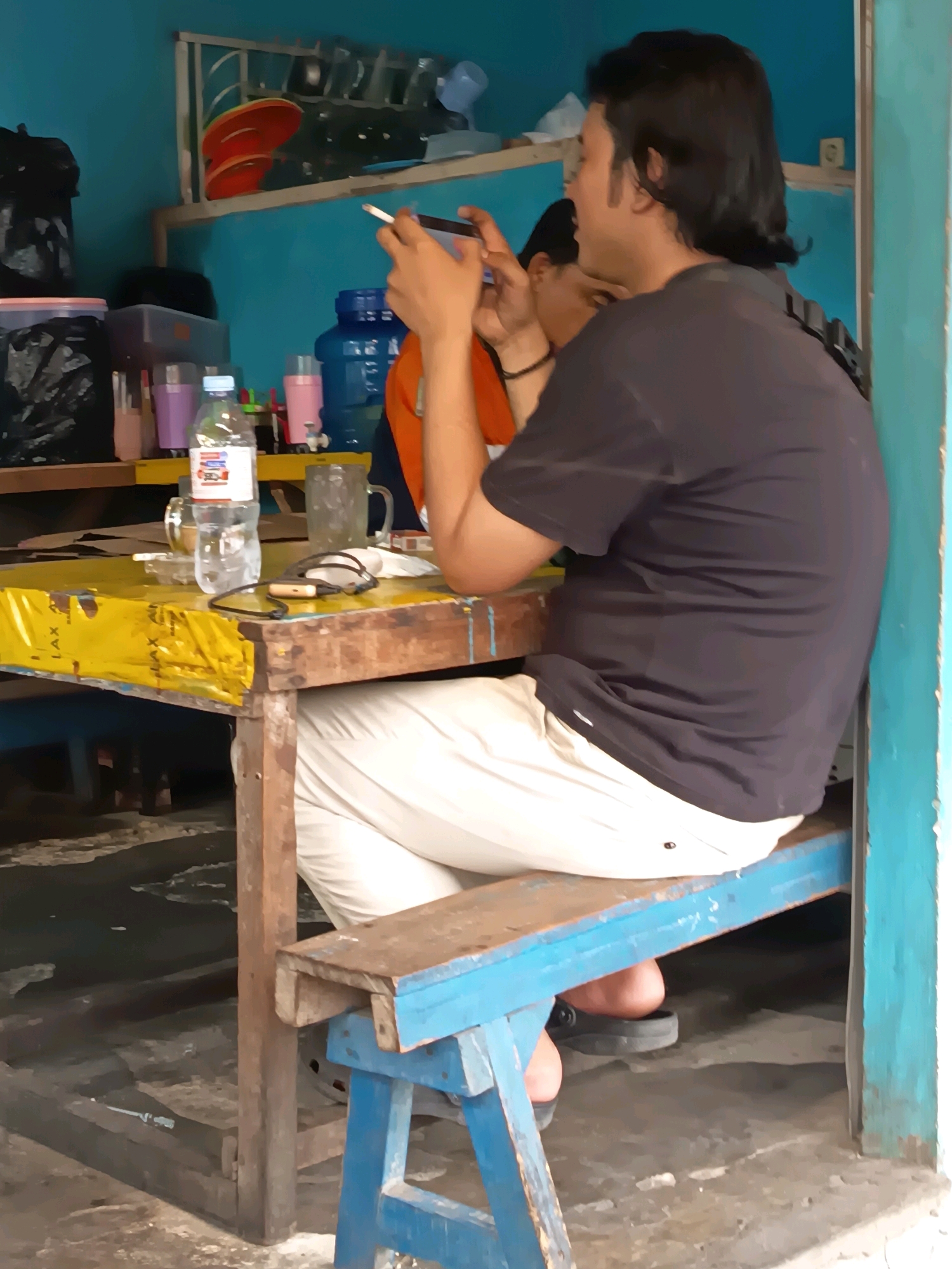 In Banjarmasin, Indonesia, a man with dark, shoulder-length hair is seated at a rustic wooden table. He is wearing a dark t-shirt and light-colored pants, and he is looking down at a mobile phone in his hands while holding a cigarette. A second person, partially visible, is seated to his left and is also looking at a phone. The table is covered with a bright yellow plastic sheet and displays a plastic water bottle, a glass, and a small ashtray with a cigarette. In the background, shelves are stocked with various containers, including large blue water jugs and smaller plastic cups, and red plates are stacked on one shelf. The walls are painted a vibrant blue. The overall environment suggests a simple, open-air eatery or roadside stall.