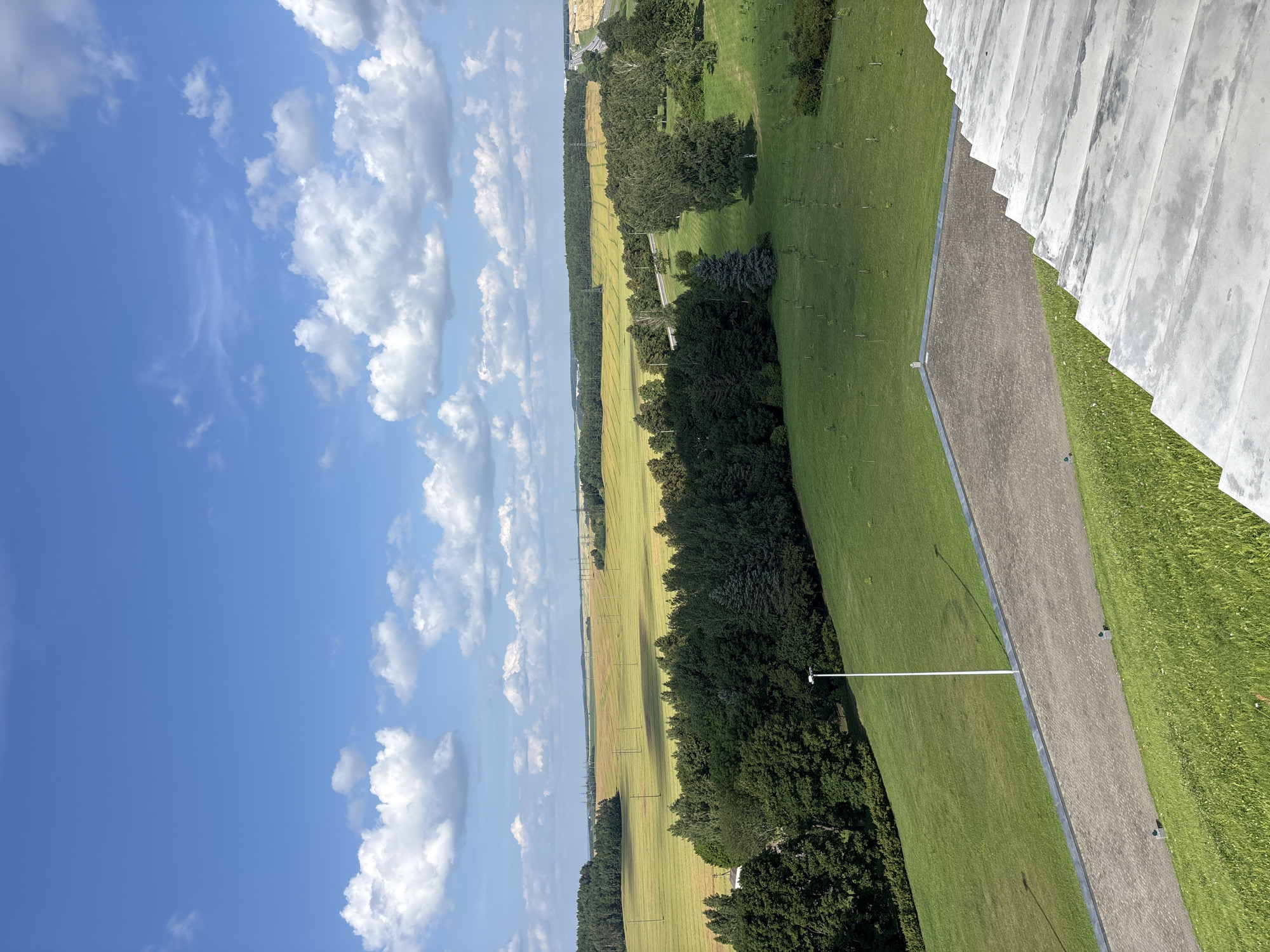 An aerial, wide-angle view of a rural landscape under a partly cloudy blue sky. In the foreground, a concrete structure with visible steps extends into the lower right of the frame, bordering a neatly mown grassy slope. A paved pathway, constructed with interlocking bricks and lined with a dark stone border, runs diagonally across the mid-ground. A tall, white lamppost stands on the grassy area adjacent to the pathway. Beyond the pathway, a dense line of dark green trees forms a transition to a large, golden-yellow field of crops, likely wheat or barley, stretching towards the horizon. In the distance, a faint treeline is visible, punctuated by power line towers. The overall scene depicts a serene, cultivated rural environment in the Ozyaritska-Slabadski rural council, Belarus.
