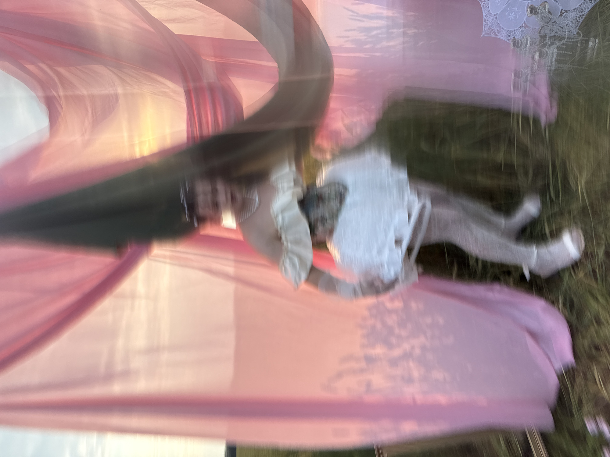 A person in a white, layered dress and sheer, patterned stockings is standing in a field with tall grass, partially obscured by pink and green draped fabric. The person is wearing a pearl necklace and a corset. A brass candelabra and a white lace umbrella are visible in the foreground. The scene is illuminated by the warm light of what appears to be a sunset. The image is blurred, suggesting motion or a long exposure. The location is the city of Malyi Bukor, Russia.