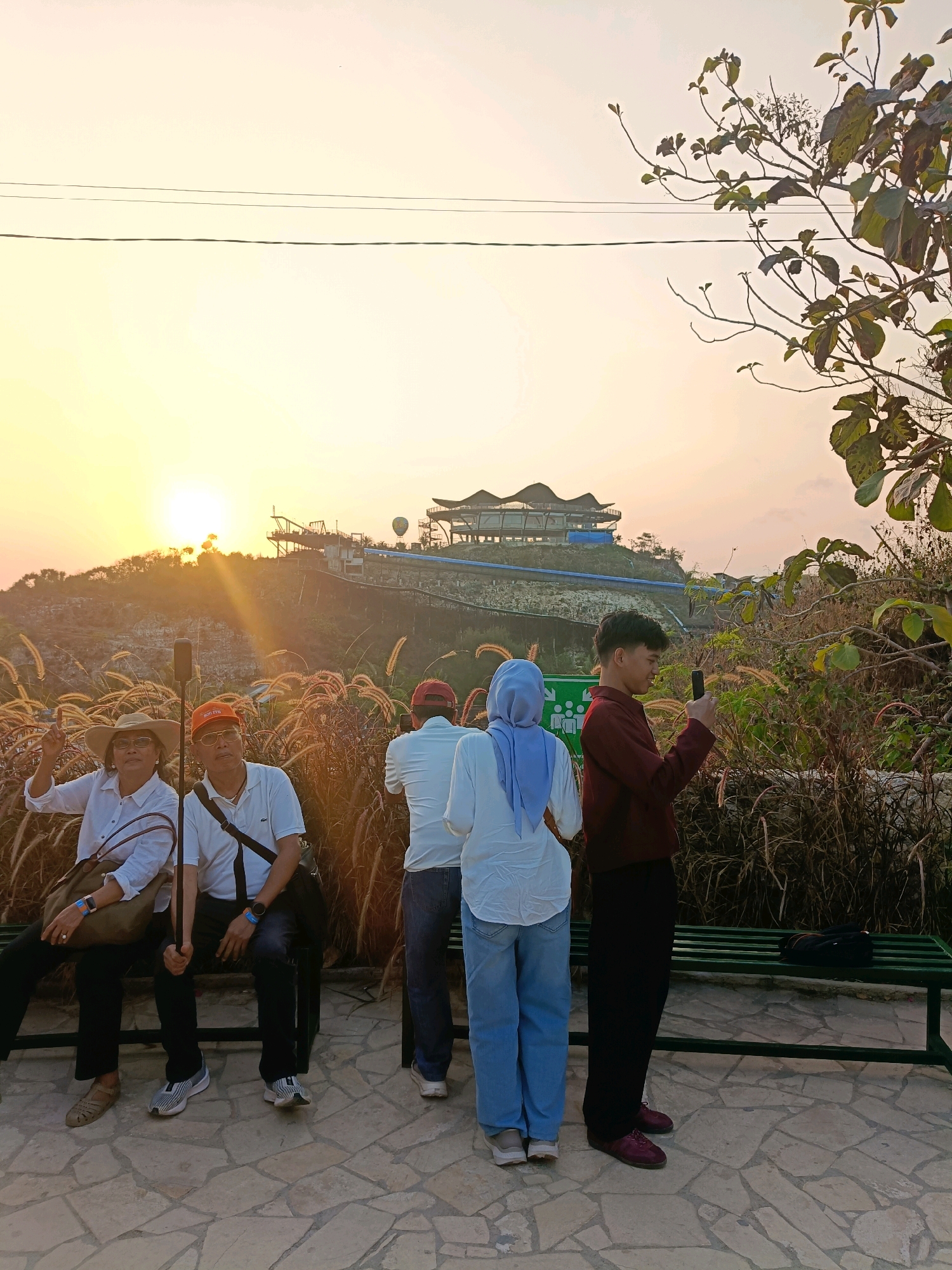 In Tanjungsari, Indonesia, at sunset, a group of people are gathered outdoors, possibly at a tourist attraction. The scene is bathed in the warm glow of the setting sun, which casts long shadows and illuminates the landscape. In the background, a multi-level structure with a distinctive roofline sits atop a hill, suggesting a man-made attraction such as a viewpoint or amusement park. Hot air balloons are visible in the sky near the structure.
In the foreground, several individuals are present. Two people are seated on a green bench. The person on the left, wearing a wide-brimmed hat and glasses, is looking towards the camera and making a peace sign. They are dressed in a white shirt and dark pants. Beside them, another person wearing an orange cap and white polo shirt is also seated. A third person, dressed in a white shirt and blue jeans, stands next to them, facing away from the camera. To the right, a young man in a maroon jacket and dark pants is standing and looking at his phone, which he is holding up as if taking a picture. Another person, wearing a light blue headscarf and white top, stands beside him. The ground is paved with irregular stone tiles. The area is also filled with tall, dry grass or reeds, adding a natural element to the scene. Power lines are visible stretching across the upper portion of the image against the pale sky.