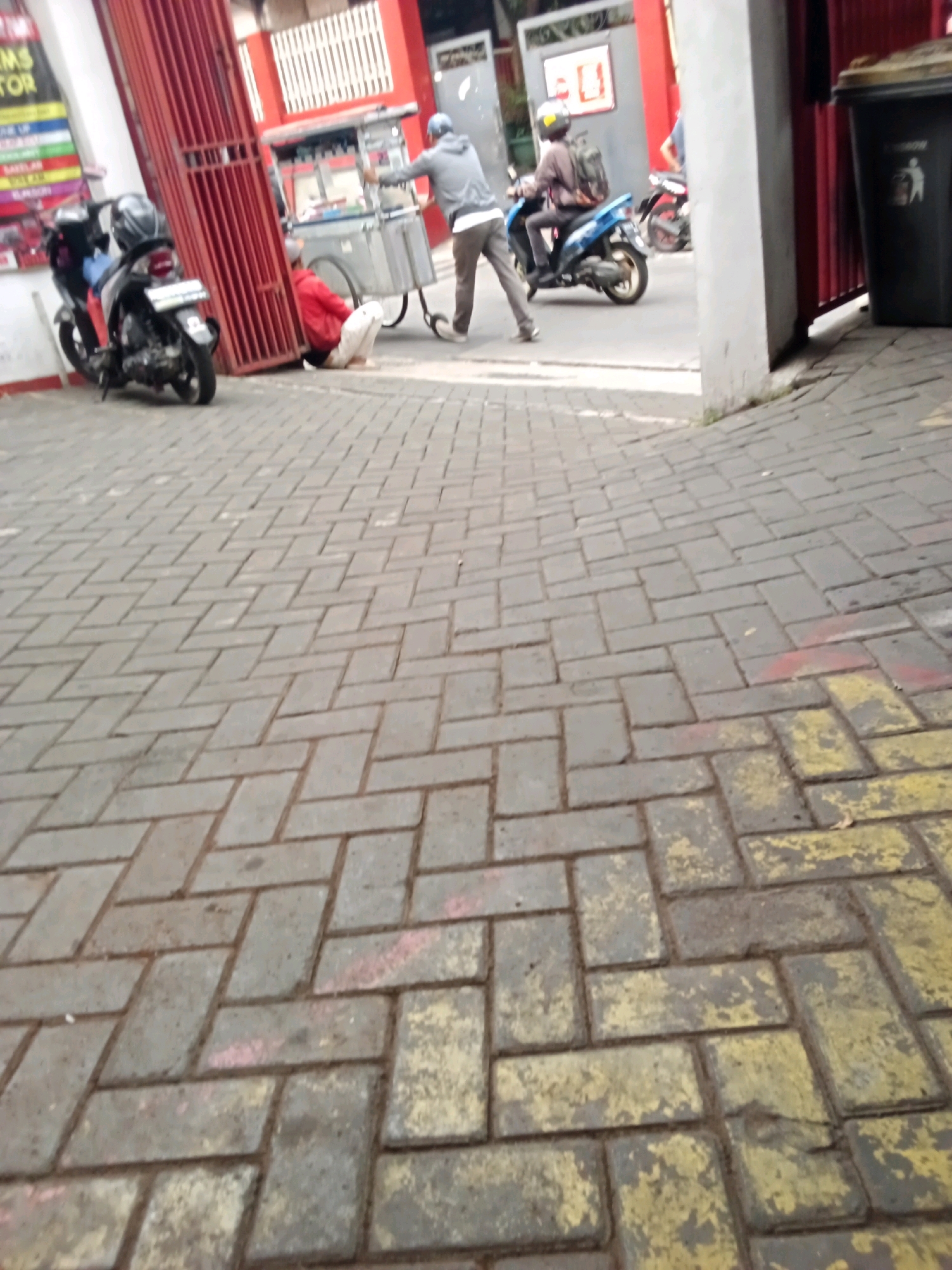 The image depicts a street scene in Bandung City, Indonesia, characterized by a paved walkway constructed from interlocking rectangular bricks. In the background, a red metal gate is prominent, with a person in a red jacket crouching near its base. Beside them, a mobile food cart with a metal frame and a white top is positioned. To the left of the cart, a person in a gray hooded sweatshirt and dark pants is pushing a smaller cart on wheels. A black scooter is parked to the left of the gate. Further down the street, a blue scooter is being ridden by a person wearing a backpack and a helmet. A dark gray trash bin with a yellow lid is situated to the right of the red gate. The walkway has patches of yellow and red markings on some of the bricks.