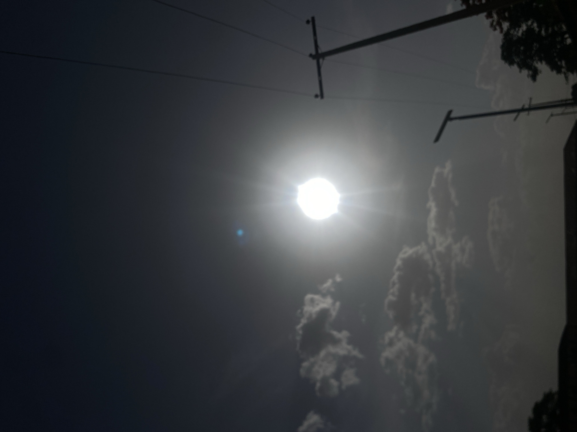 The image shows a bright sun in a hazy sky, obscured by thin clouds. Power lines and poles are visible in the upper portion of the image. Based on the location of Tudun Wada, Nigeria, the presence of power lines suggests a developed area. The hazy sky could indicate dust or pollution, common in some urban environments.