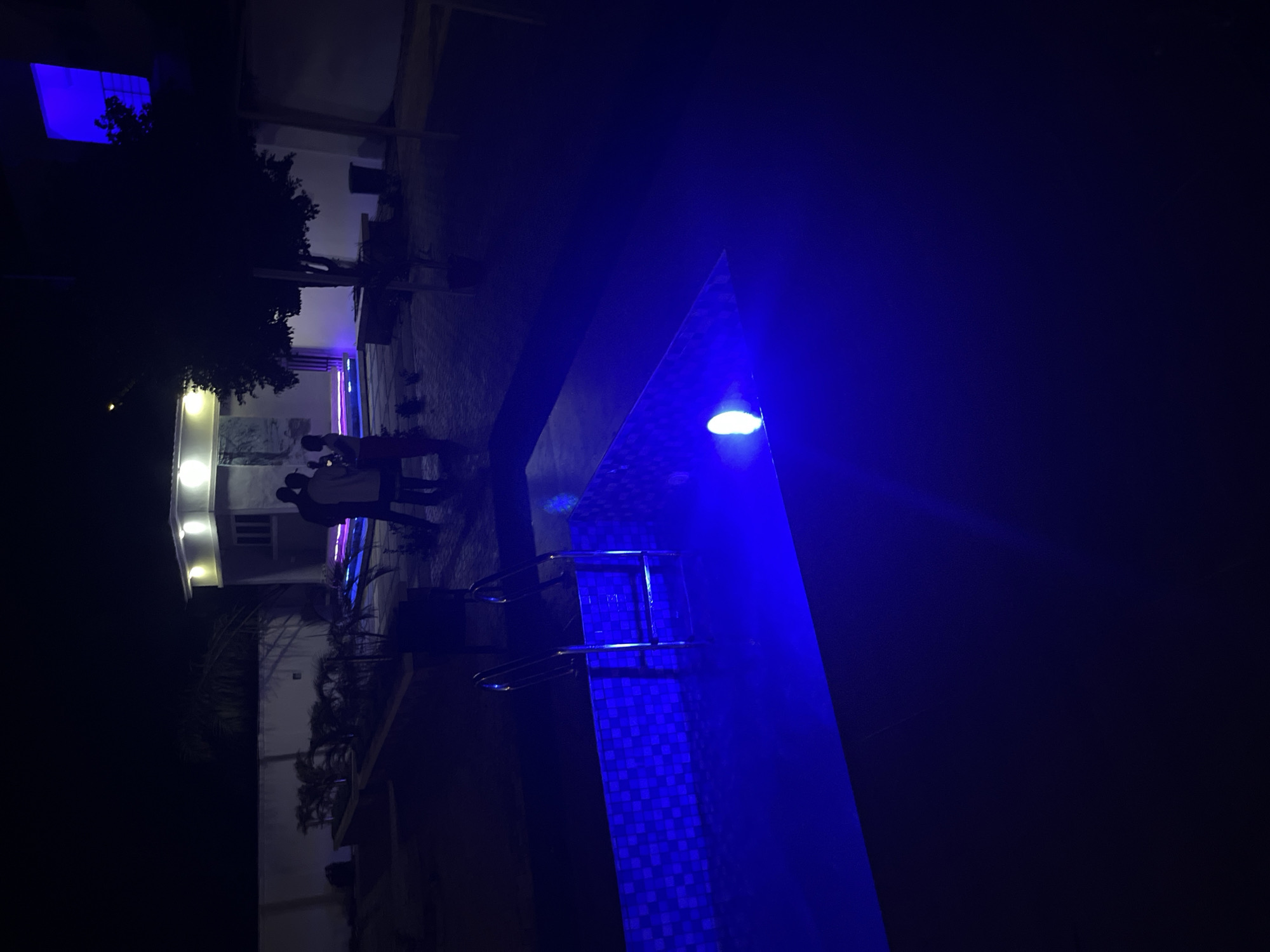 The image depicts a nighttime scene at a residential property in Kason, Nigeria, featuring a swimming pool illuminated by blue underwater lights. The pool is rectangular with tiled edges and a metal ladder. Adjacent to the pool, several individuals, silhouetted against the ambient lighting, are gathered. In the background, a modern building with illuminated windows and a flat roof is visible, surrounded by a walled perimeter and some vegetation. The overall ambiance suggests a social gathering or relaxation at a private residence.