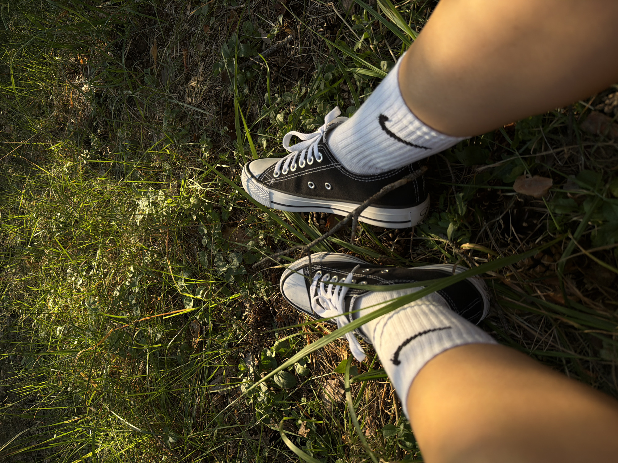 The image shows a pair of feet wearing black high-top Converse sneakers with white laces and white crew socks with a black Nike logo. The feet are positioned in the grass, with sunlight filtering through the blades. The background consists of green grass and dry leaves, suggesting an outdoor environment. The scene is captured from a first-person perspective, looking down at the feet. The location is identified as Udelnaya, Russia.