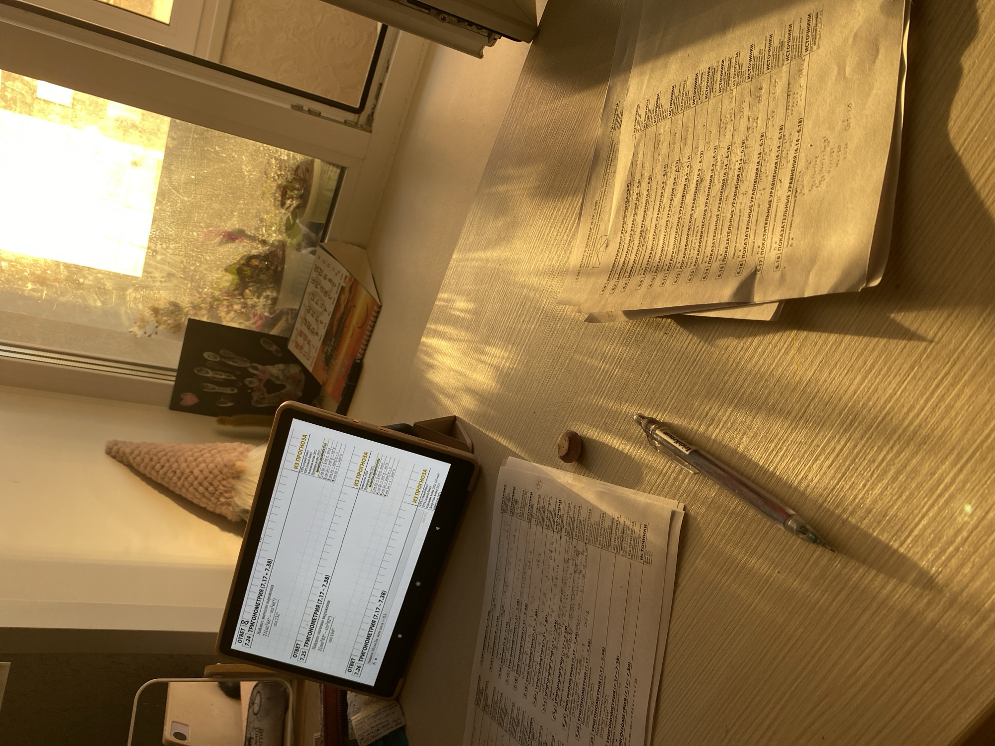 The image shows a desk illuminated by sunlight streaming through a window. On the desk, there's a tablet displaying text, several sheets of paper filled with mathematical formulas and problems in Russian, and a pen. Near the window, there are decorative items including a small figure and a calendar. The setting appears to be an indoor workspace, possibly a study area.