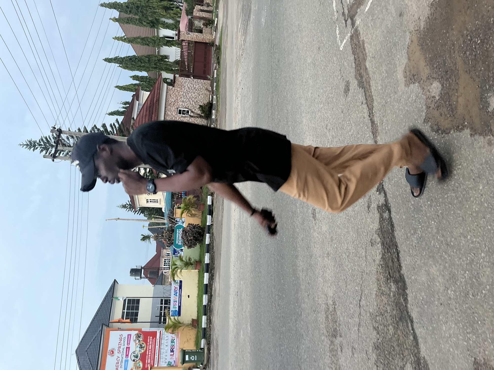 A young man wearing a black t-shirt, a black baseball cap, tan pants, and dark slide sandals is walking along a paved road in Abuja, Nigeria. He is walking from left to right in the frame, with his head turned to the left and his left hand touching his chin. He is holding a black object in his right hand. In the background, there are several buildings, including a school with signs indicating "NURSERY," "PRIMARY," and "SECONDARY SCHOOL." There are also trees and power lines visible. The sky is overcast. The road surface appears wet in places.