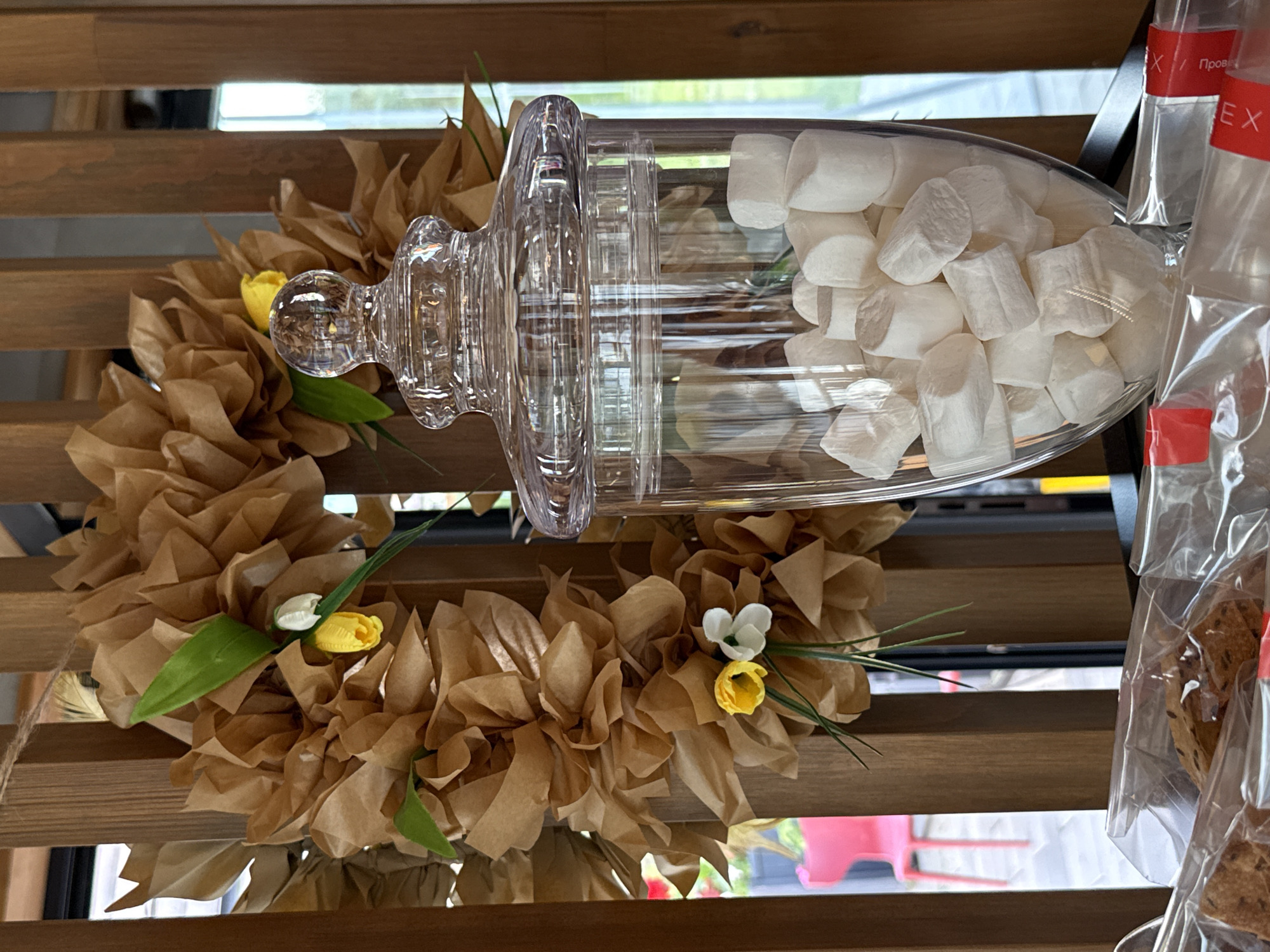 This media file shows a close-up of a decorative display, likely in a shop or cafe in Votkinsk, Russia. The main subjects are a clear glass jar filled with marshmallows, resting on a wooden shelf. Beside the jar, a wreath made of crumpled brown paper is decorated with artificial flowers, including small yellow and white tulips, and green leaves. The wreath is tied with a twine string.
In the background, another wooden shelf is visible, with some bagged items to the right of the frame. These bags appear to contain baked goods, possibly cookies or crackers, and have red labels with white text. The text on one visible label appears to be "EX" followed by Cyrillic characters, likely "Пробе". The overall scene suggests a retail environment, possibly decorated for a spring or Easter theme due to the floral elements and the soft, light colors.
The lighting suggests it is daytime, with natural light filtering in, possibly from a window. There are no people visible in the frame. The image is captured from a slightly elevated angle, looking down at the display. The focus is on the arrangement of the jar, marshmallows, and wreath, with the background elements slightly blurred.