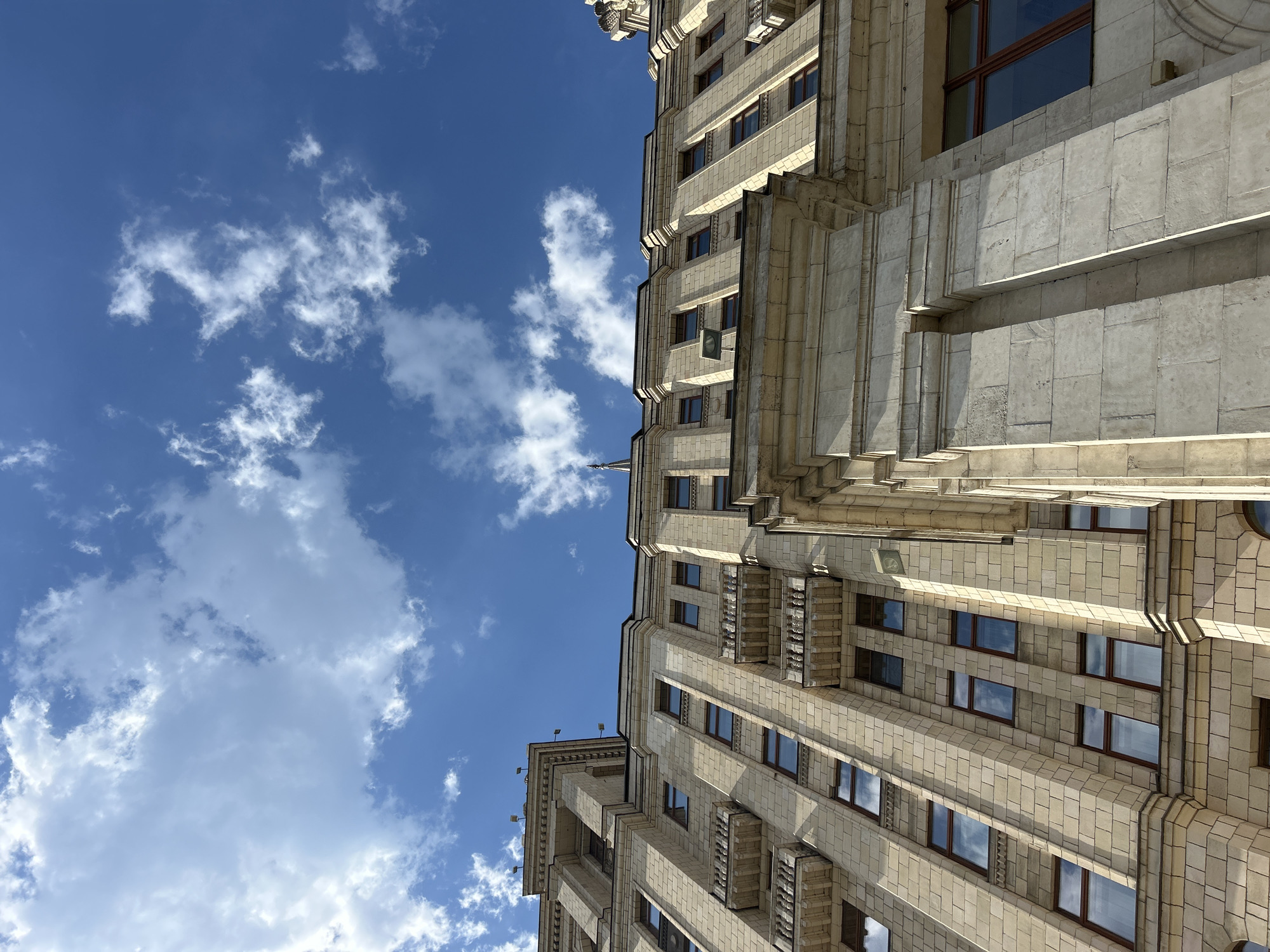 The image is a low-angle shot of a tall, light-colored building set against a bright blue sky with scattered white clouds. The building features a classical architectural style with multiple floors, many windows with dark frames, and prominent vertical architectural elements. The facade has a textured, stone-like appearance, and there are protruding balconies or architectural details at certain levels. The building is likely located in an urban area, but the exact location within the city is not identifiable from the image. There is no observable human activity in the shot.