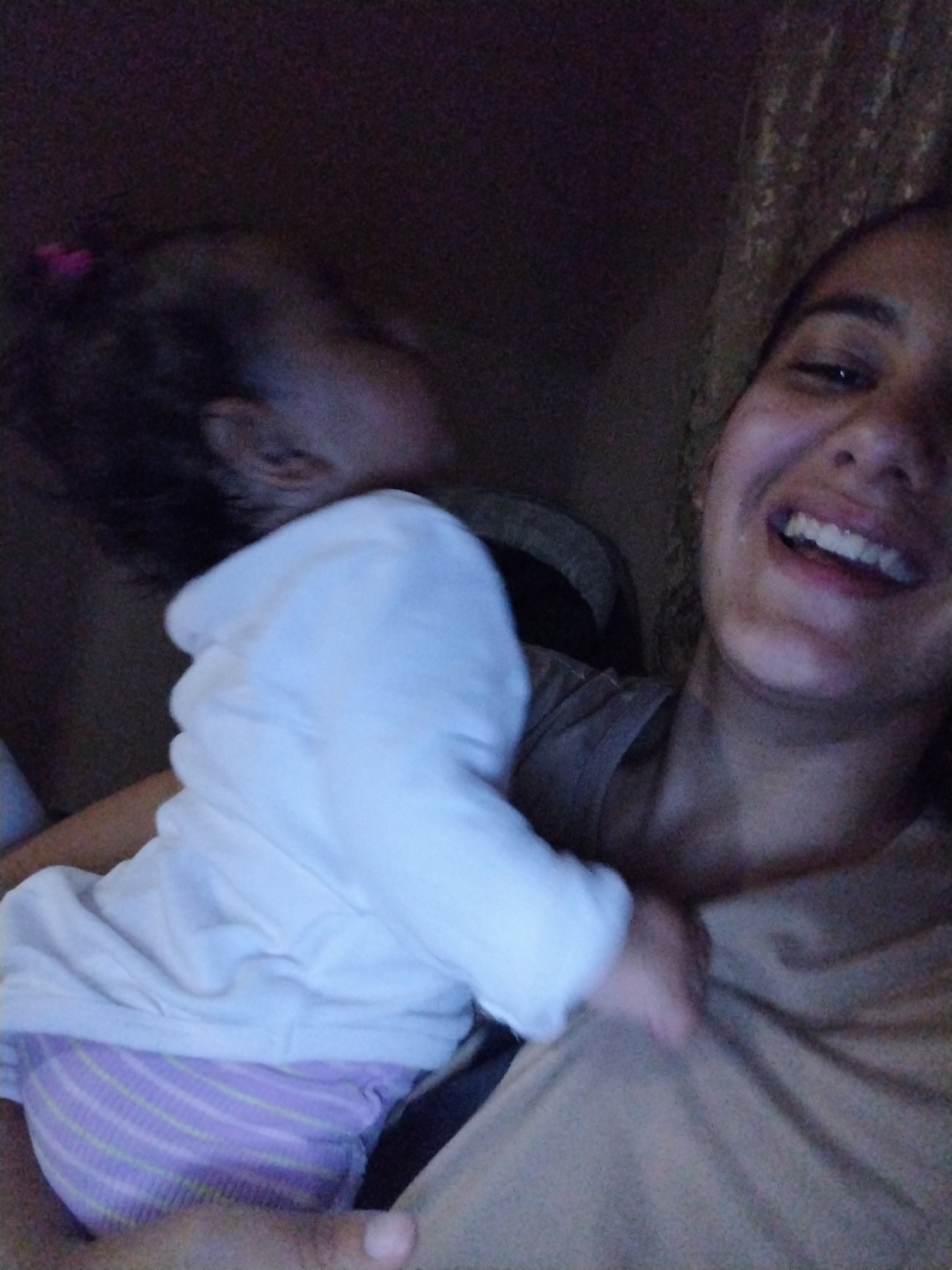 The image captures an adult female holding an infant in a dimly lit indoor setting. The adult female, positioned on the right side of the frame, has a medium complexion and dark hair. She is wearing a light brown or tan top and is looking directly towards the camera with a wide, open-mouthed smile, revealing her teeth. The infant, held close to the female's left shoulder, is primarily visible from the back and left side. It has dark hair, secured with a small pink hair tie on the left side of its head. The infant is dressed in a white long-sleeved top and a purple-and-light-green striped diaper or bottoms. Its head is turned to its left, facing away from the camera. The background is dark and largely indistinct, suggesting low ambient light. A faint, dark patterned texture is discernible on the far right. The scene is located in Unknown, Unknown.