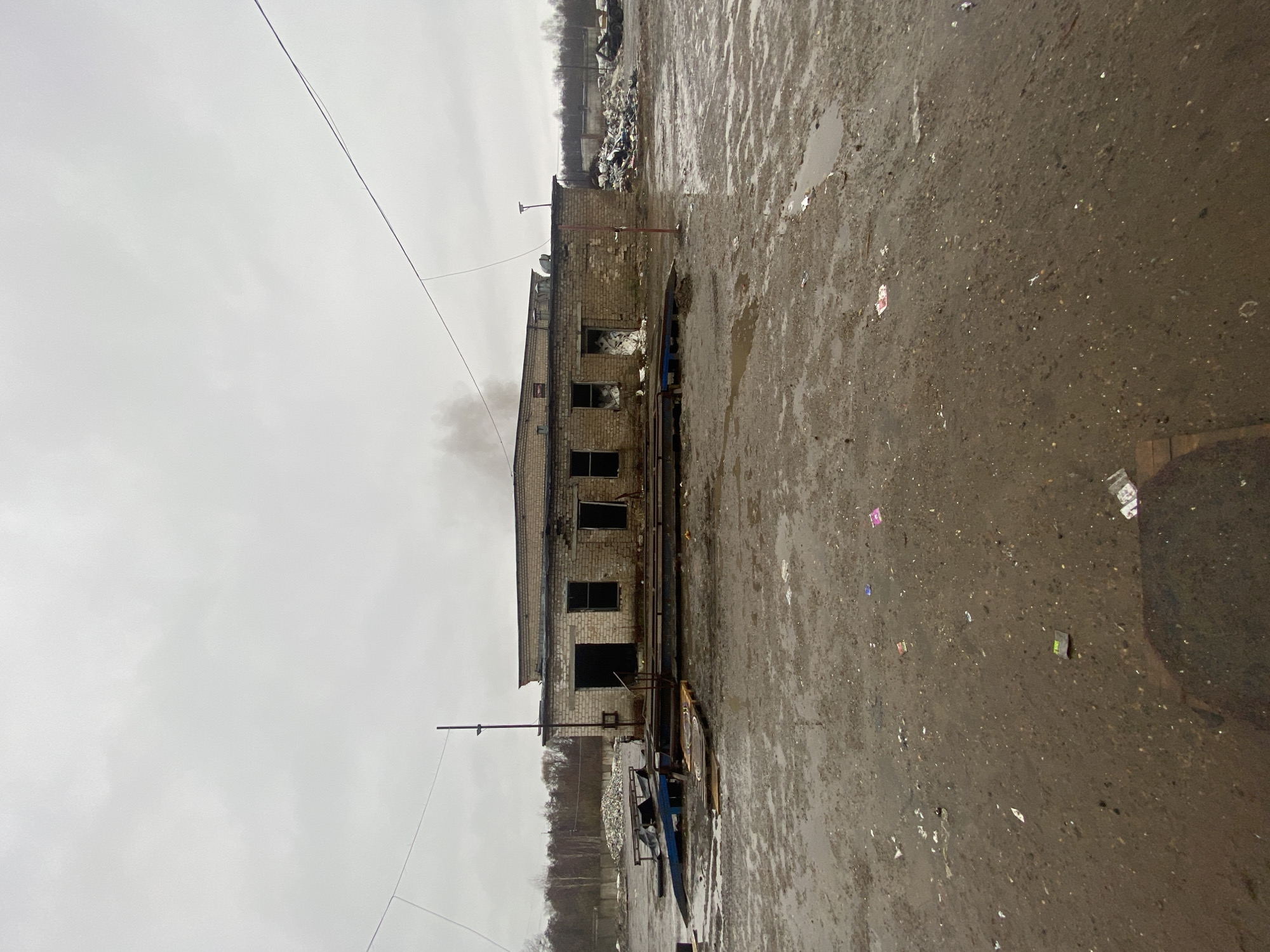 The image shows a dilapidated brick building with broken windows and a collapsed roofline. Smoke is rising from the building. The ground in front of the building is a wet, asphalt-like surface littered with debris and puddles. In the background is a pile of debris, possibly trash or rubble, and some bare trees. Above is an overcast sky. The location is Ol'khovskoye, Russia. There are no people visible in the image.