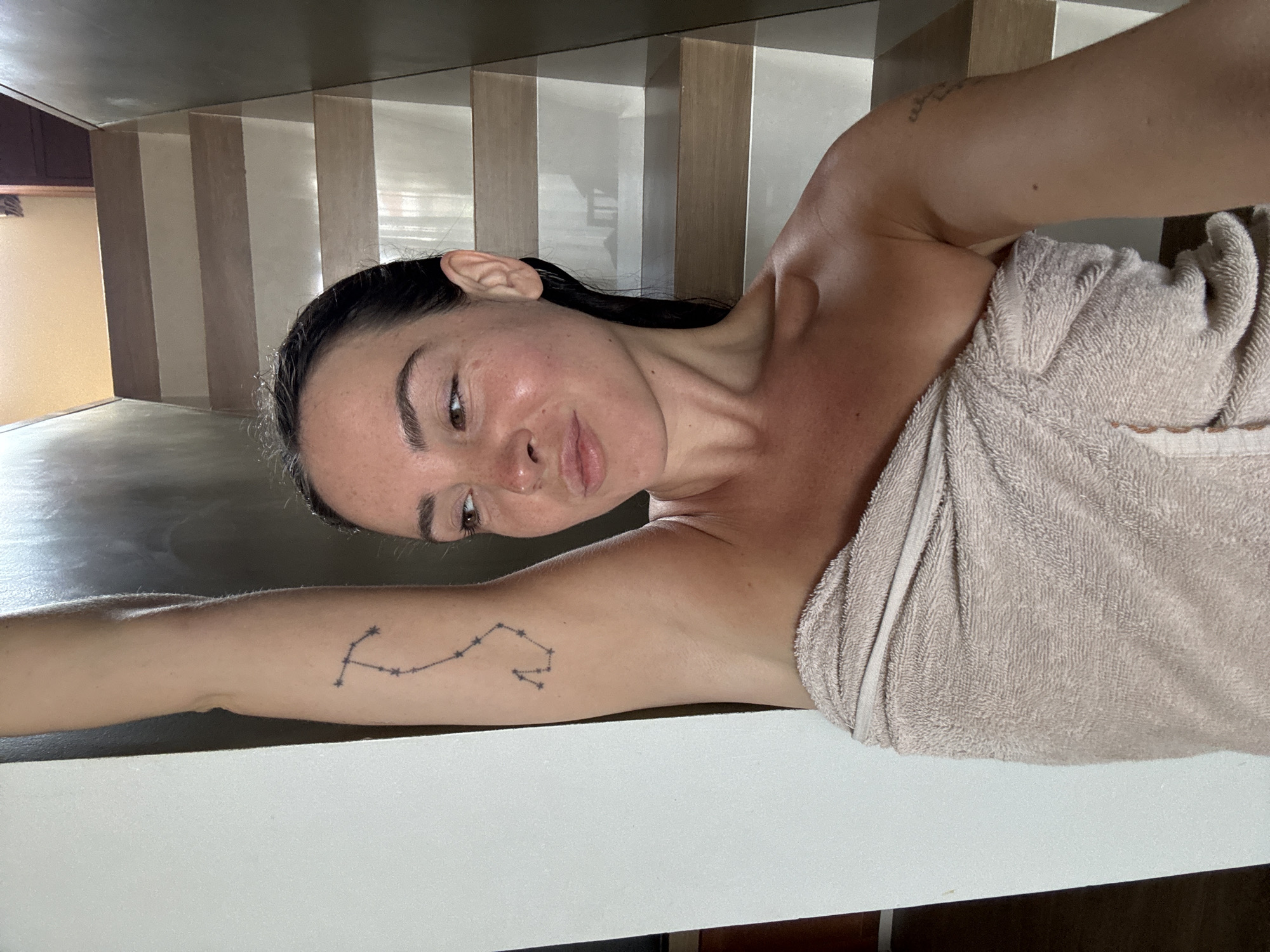 A young woman is taking a selfie in what appears to be a stairwell. She is wearing a beige towel wrapped around her body, and her left arm is raised. She has a tattoo of a constellation on her upper left arm. The stairs are made of wood and are illuminated by natural light. The background is a wall that is partially white and partially dark. The woman is looking to her right and her lips are slightly parted.