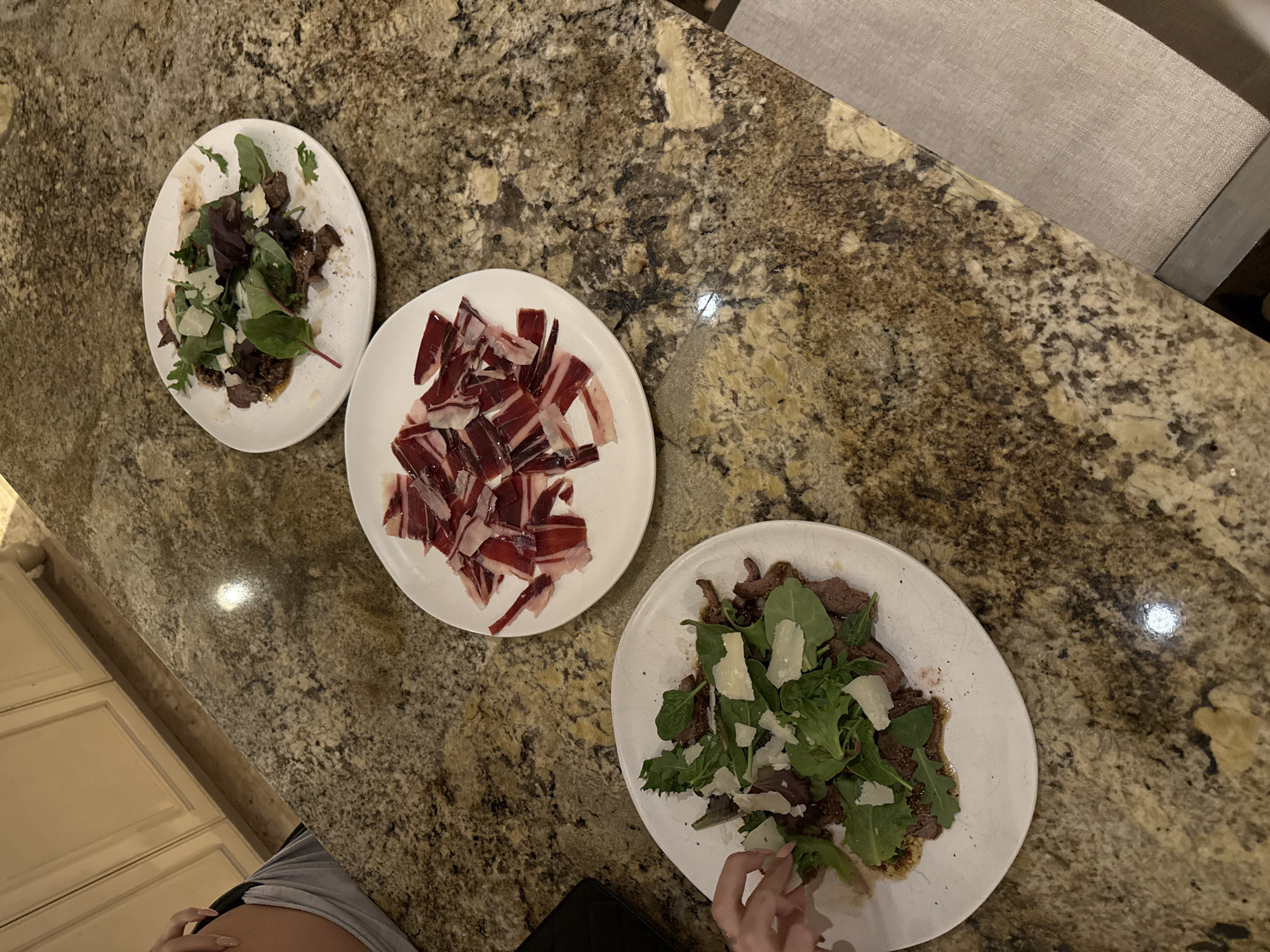 The image depicts three white plates on a granite countertop. One plate contains what appears to be thinly sliced ham, while the other two plates contain a salad mixture of greens, meat, and shavings of a light-colored cheese. A hand is visible near one of the salad plates, touching the greens. The environment suggests an indoor dining area, with part of a chair and a glimpse of cabinetry visible. There's no explicit information within the image itself to confirm a specific location in Miami Beach or elsewhere.