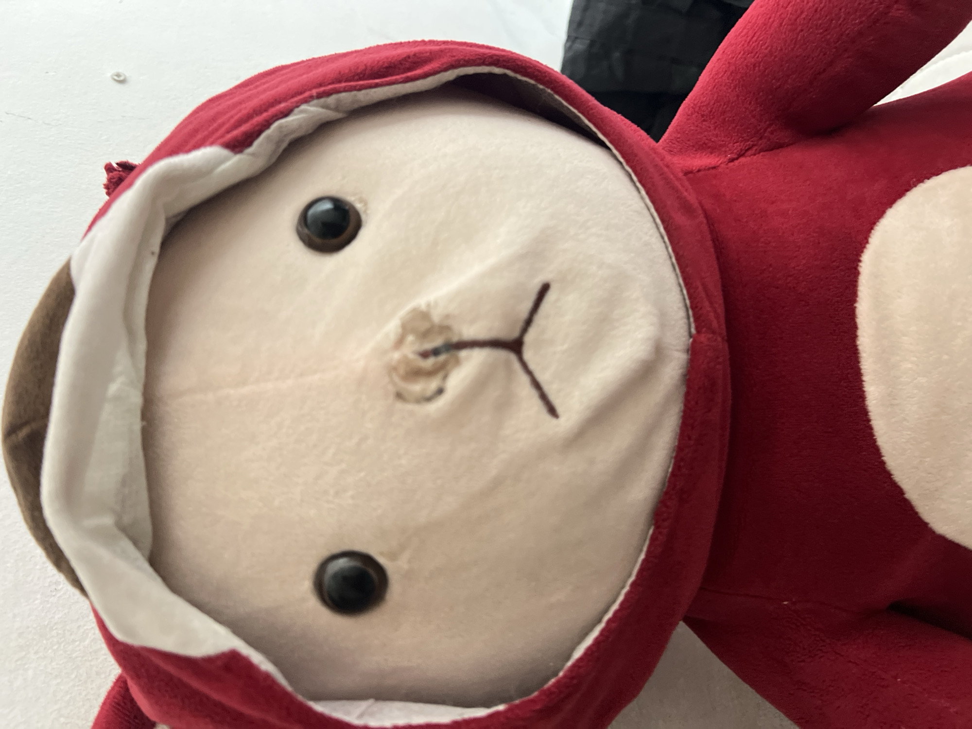 A close-up, eye-level, indoor shot shows the face of a plush toy bear wearing a red hooded costume. The bear has light brown fur on its head and face, dark brown eyes, and embroidered facial features, including a small nose and a mouth shaped like an upside-down 'T'. The hood is red with a white lining and brown ears. The toy is positioned against a white wall. The image was taken at the Denpasar, Indonesia location.