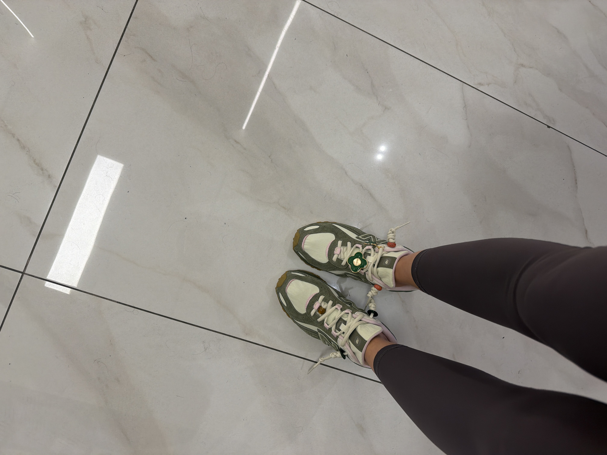 A high-angle indoor shot shows a person wearing grey leggings and sneakers standing on a white marble tile floor with grey grout lines. The sneakers are a light grey-green and off-white color with light beige laces. The floor is illuminated by a linear reflection of light. The location is within Yerevan, Armenia. No other people or objects are visible.