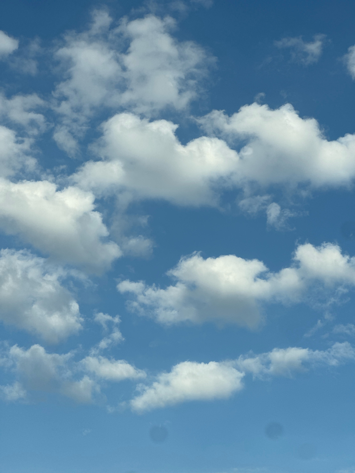 The image is a close-up shot of a bright blue sky with scattered white clouds. The clouds vary in size and density, with some appearing fluffy and well-defined, while others are wispy and less opaque. The overall impression is of a clear, sunny day. The scene is devoid of any man-made structures or natural landscapes, focusing solely on the atmospheric conditions. There are no people, animals, or identifiable objects present. The image captures a section of the sky that could be observed from anywhere, including the city of Mesa Geitonia, Cyprus, on a day with fair weather. The dark circular spots in the lower portion of the image are artifacts, possibly from the lens or sensor of the camera.