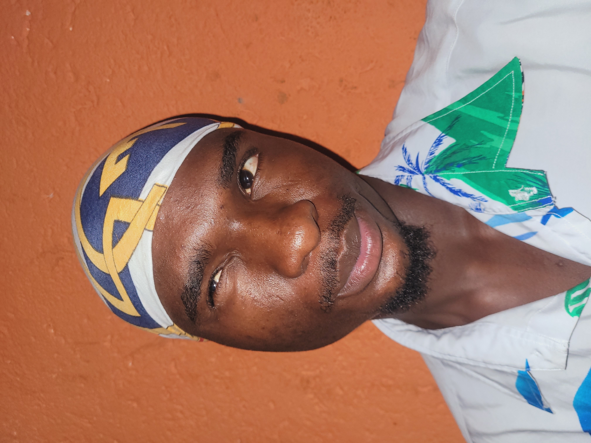 A young Black male with a light complexion is photographed from the chest up, looking to his right with his left eye slightly squinted. He is wearing a white button-down shirt with a tropical print of green palm trees and blue abstract shapes. A bandana with a pattern of blue, yellow, and white geometric shapes covers his head. The background is a textured orange wall. The image appears to have been taken indoors, possibly in a residential setting, given the close-up framing and the backdrop. There are no other individuals or objects of note visible in the frame. The lighting suggests a single light source from the front, creating subtle shadows on the subject's face.
