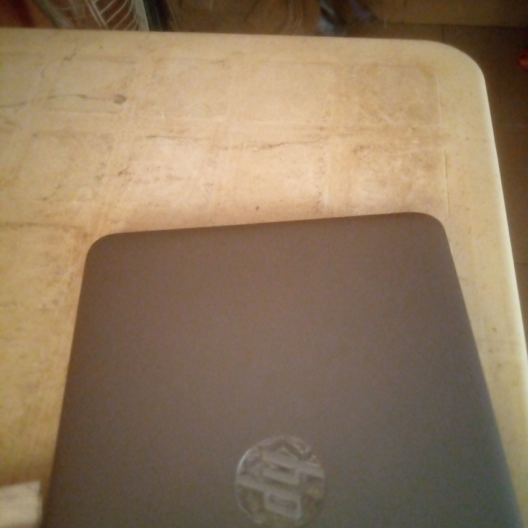 The image shows a closed laptop with a silver logo on its dark gray lid, resting on a beige surface. The surface appears to be a table with a textured pattern. The background is out of focus and shows some indistinct shapes and colors, suggesting an indoor setting. The lighting is dim.