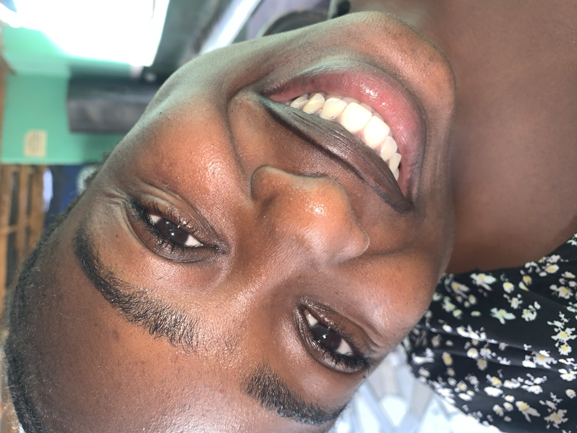 An overhead close-up shot captures a person, likely female, with dark skin, smiling broadly towards the camera. The image is oriented sideways, with the person's head tilted to the right. The prominent feature is a wide, joyful smile revealing healthy, white teeth and full, pink lips. Her dark eyes are open and reflect light, and her eyebrows are dark and well-defined. The skin on her face appears moisturized with a slight sheen on the forehead, nose, and cheeks.
The subject is wearing a black garment with a white and yellow floral pattern, visible on the right side of the frame. The background is out of focus but shows an indoor setting. To the left, a turquoise or mint green wall is visible, with a dark, rectangular object, possibly an air conditioning unit, mounted on it. A faint light-colored square shape is also present on the green wall further down. The overall lighting is bright, suggesting a well-lit interior. No visible text is present in the image. The provided context indicates the location is New Stadium, Nigeria.