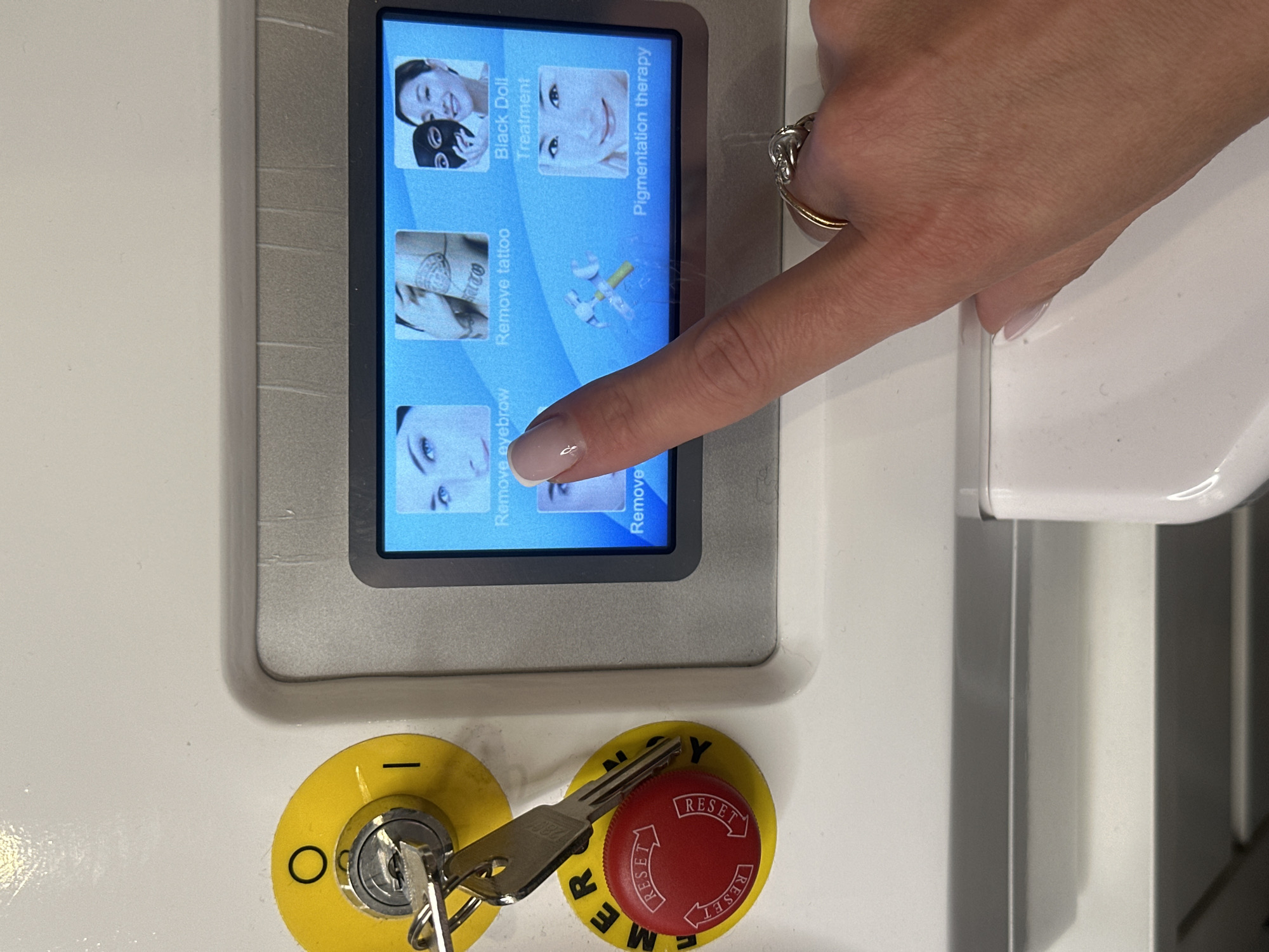 A close-up photograph shows a person's hand touching the screen of a machine, possibly a cosmetic or medical device. The screen displays several options with images and text, including "Black Doll Treatment," "Pigmentation therapy," "Remove tattoo," and "Remove eyebrow." The person's hand has a ring on one of the fingers and a neatly manicured nail. Below the screen, there are two yellow emergency stop buttons, one with a key inserted. The environment is clean and sterile, suggesting a professional clinic or salon setting. The photograph does not contain any information to suggest Riga, Latvia.