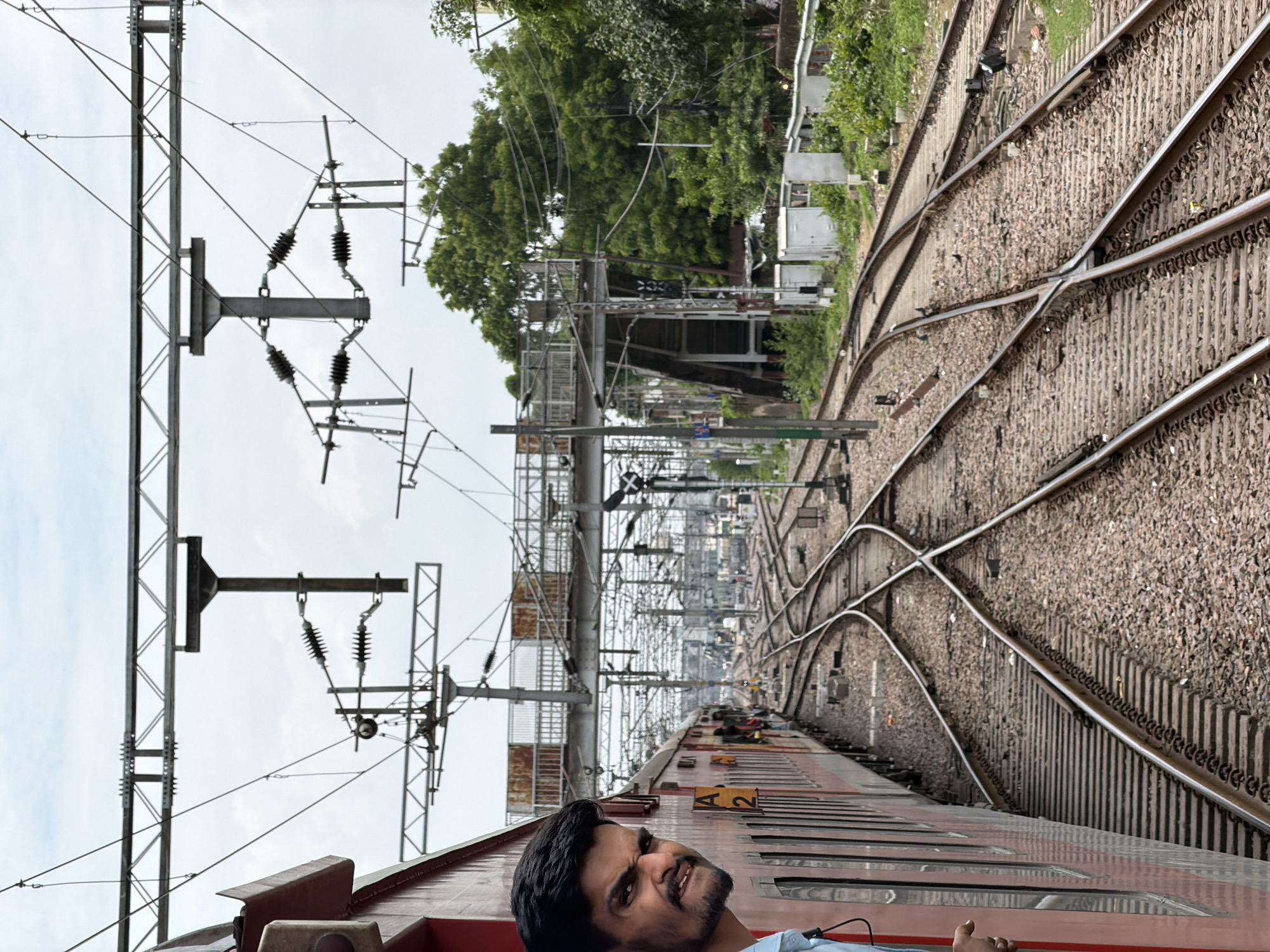 The image captures a view of a train track junction in Kanpur, India. A person is leaning out of the window of a red train, making a facial expression at the camera. The train is positioned on the left side of the frame, while the right side shows multiple converging train tracks. Overhead, electrical wires and support structures are visible against a cloudy sky. In the background, there are trees, a pedestrian bridge, and some small buildings. The overall scene depicts a busy railway environment.