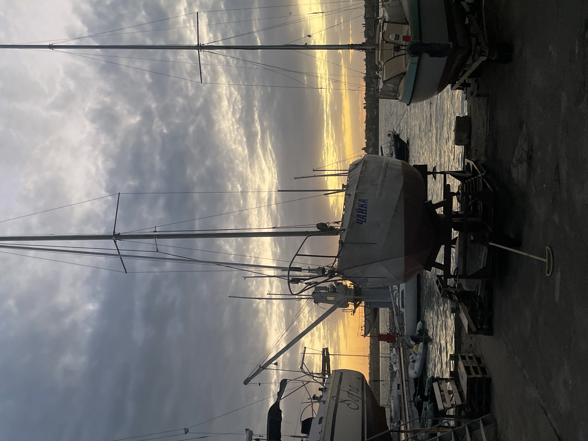 The image depicts a harbor scene at sunset, possibly in the city of Unknown, Unknown. Several sailboats are visible, some in the water and others on stands ashore. The boat in the foreground is labeled "ЧАЙКА" in Cyrillic script. Another boat in the lower portion of the image has the name "Sofia" written on it. The sky is filled with dramatic, dark clouds and the sun is casting a golden light across the water and harbor. A variety of marina equipment, including scaffolding and wooden pallets, can be seen on the pier.