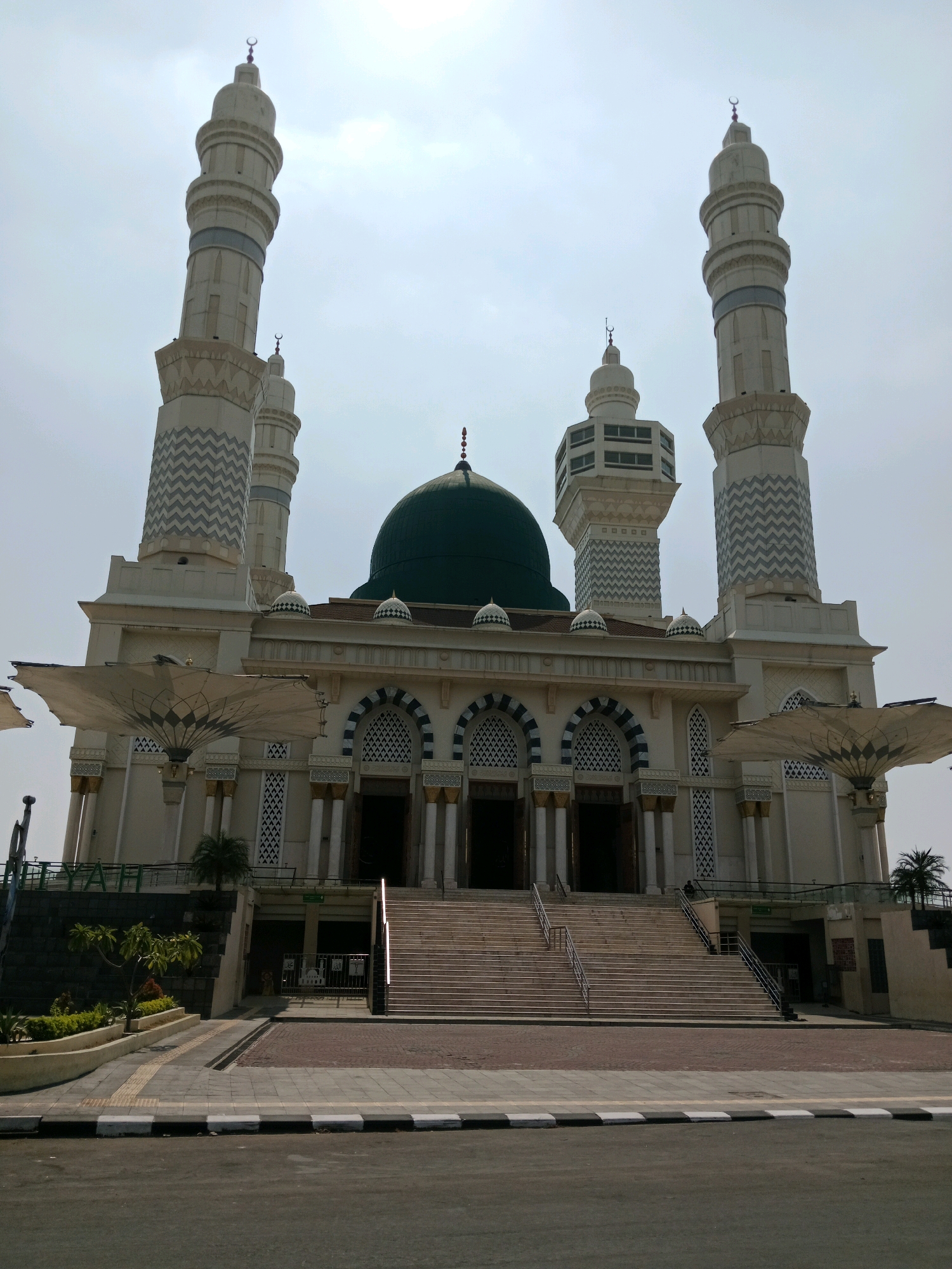 The image shows the façade of the Al-Ansar Mosque, located in Karanganyar, Indonesia. The mosque is characterized by its prominent green dome and four tall minarets, two on each side of the central structure. The minarets are white with decorative grey zigzag patterns and topped with crescent moons. The mosque's main building features large arched windows and entrances, flanked by columns. Two large, retractable sunshades, reminiscent of umbrellas, are deployed on either side of the entrance steps. A wide staircase leads up to the mosque's main level. The foreground shows a paved area with a road bordering it. The sky is overcast and bright.