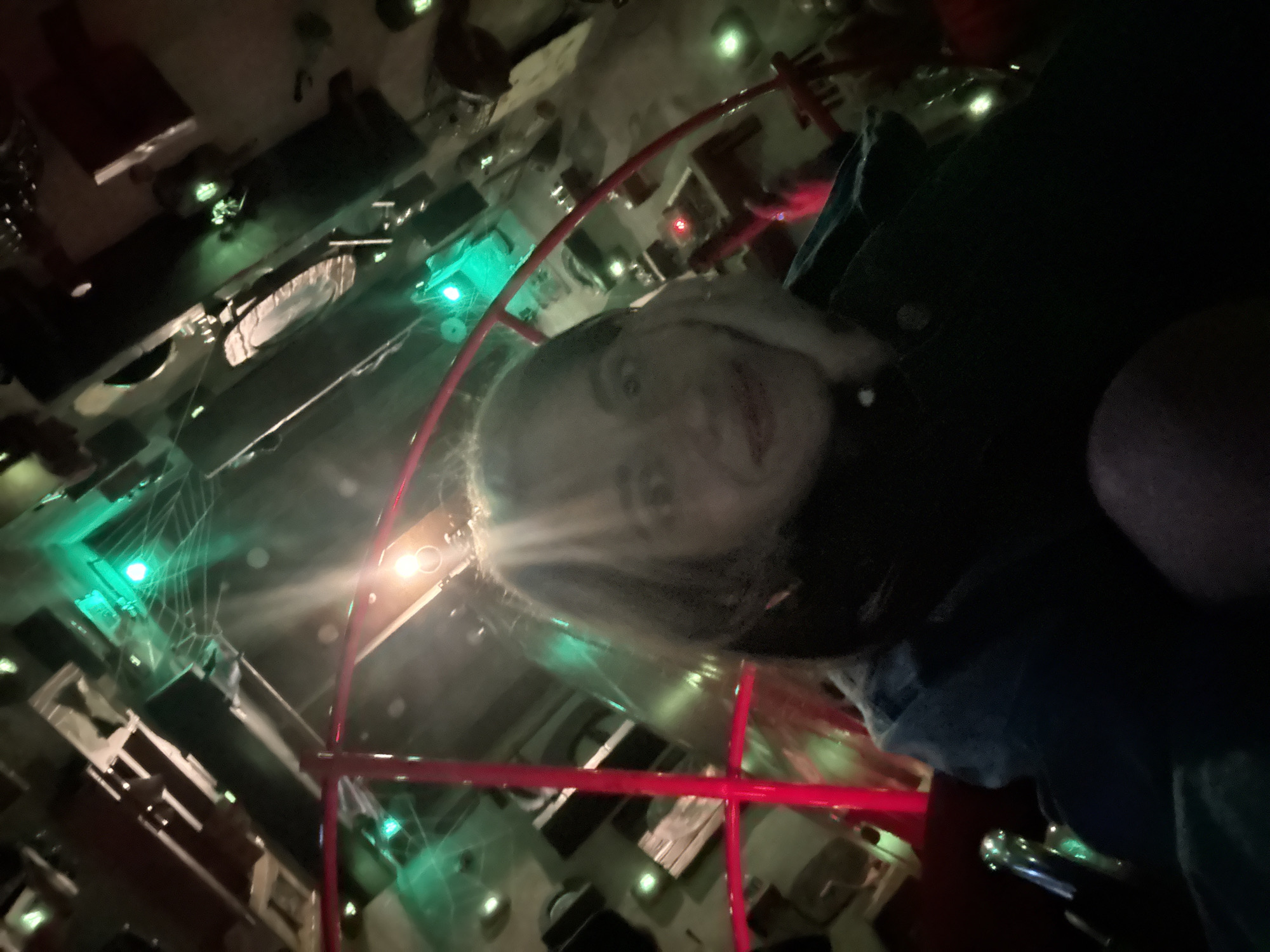 A low-angle indoor shot depicts a fair-skinned person with blond hair in the foreground, partially obscured by a red, metal structure. The person is wearing dark clothing with a visible blue denim section. In the background, an unusual interior with dark, geometric shapes lit by green and white lights suggests a stylized or themed environment. Based on visual cues, this might be a bar or entertainment space with unique decor. While the image has some recognizable elements, the specific location in Moscow, Russia is not discernible solely from the image's content.