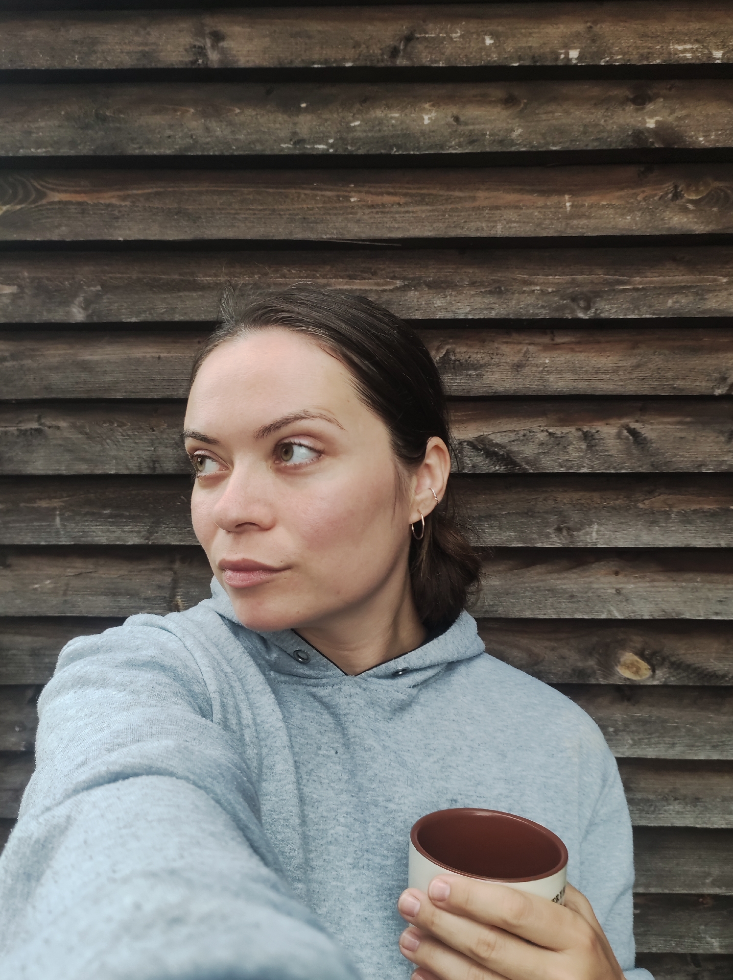 A young adult female is depicted in Фоки, Russia, positioned in front of a weathered dark wooden plank wall. She wears a light gray hooded sweatshirt. Her dark hair is pulled back. The visible ear features two small hoop earrings. She holds a ceramic mug with a white exterior and brown interior in her right hand. Her head is turned slightly to the viewer's left, with her eyes also directed in that direction.