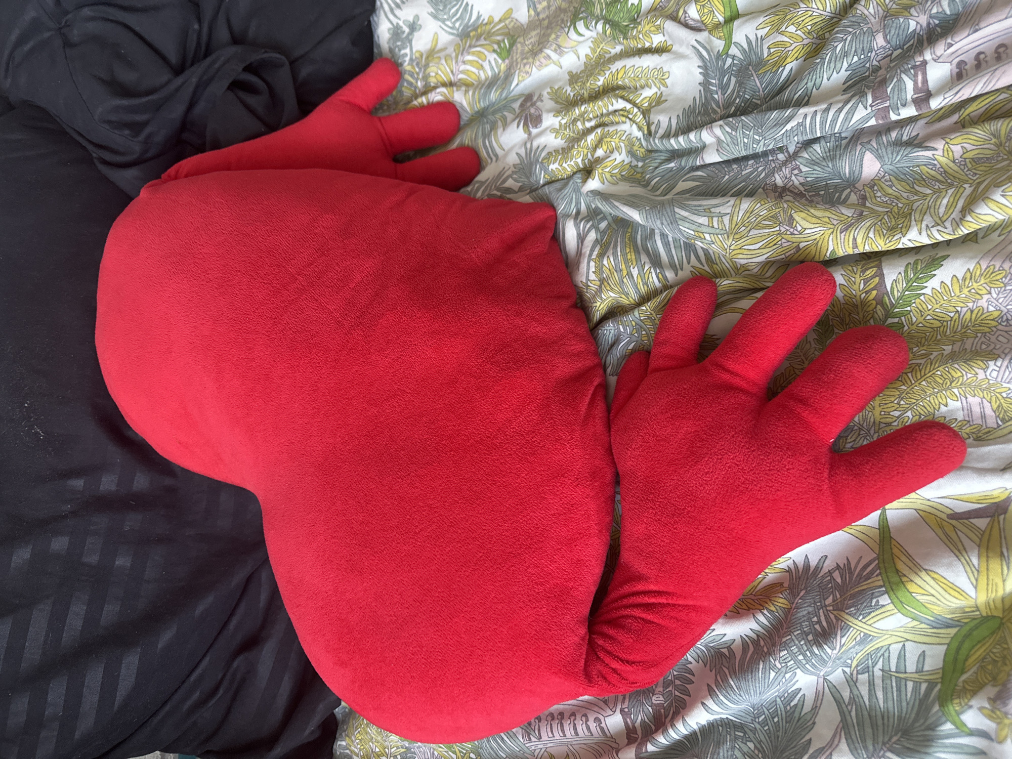 A red, plush pillow with the shape of a heart and attached hands is lying on a bed. The pillow is a vibrant red and has a soft, fuzzy texture. It is positioned diagonally across the frame, with its pointed end towards the lower left and its broader base towards the upper right. Two of the pillow's hands are visible, one extending to the left and the other to the right. These hands are also red and have a similar plush texture. The pillow is resting on a bed with a patterned duvet cover. The duvet features a botanical print with stylized leaves and trees in shades of green, yellow, and brown, set against a white background. To the left of the pillow, a section of a black blanket or sheet is visible, appearing to be made of a slightly shiny material with subtle vertical stripes. The lighting suggests it is daytime, with natural light illuminating the scene. The setting appears to be a bedroom or a living room area where the bed is located. No people are visible in the image, and no text is present.