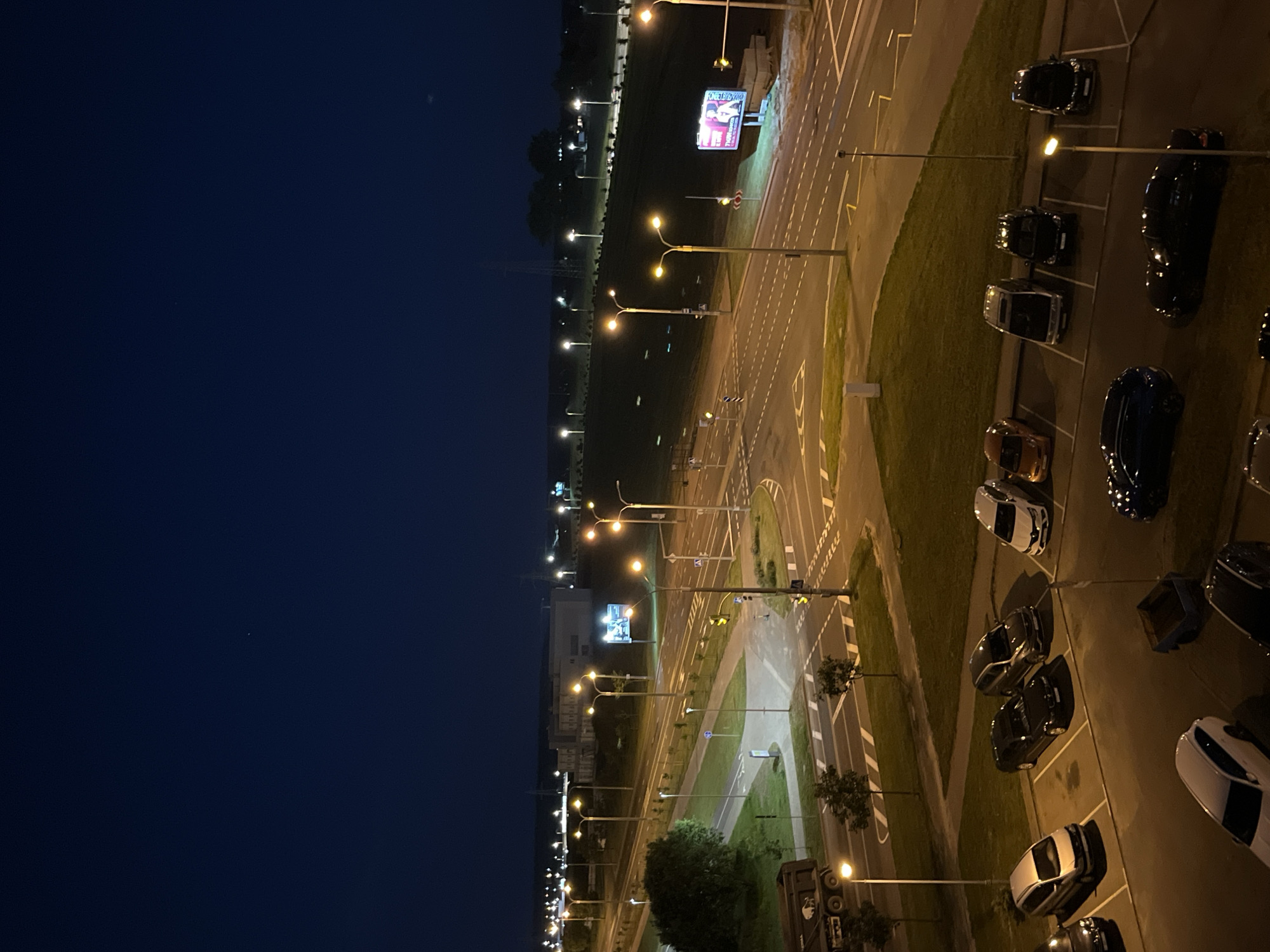 This is an aerial night-time photograph taken in Minsk, Belarus. The scene depicts a well-lit parking lot adjacent to a road. Multiple cars, mostly dark-colored, are parked in designated spots. Streetlights illuminate the area, casting a warm glow on the asphalt and surrounding grass. The sky is a deep, dark blue, suggesting it is nighttime, and there are no visible indications of weather conditions.
The parking lot appears to be part of a larger commercial or public area, as a billboard with advertisements is visible in the distance, illuminated by spotlights. The road itself has clear lane markings and traffic signals, indicating ongoing, albeit minimal, activity. The overall impression is one of quietude and order, typical of a parking area after business hours. No people are visible in the image.
The photograph is oriented vertically, with the sky at the top and the parking lot and road extending downwards. The perspective is from a high vantage point, looking down onto the scene. The lighting creates distinct shadows and highlights, accentuating the forms of the vehicles and the textures of the surfaces.