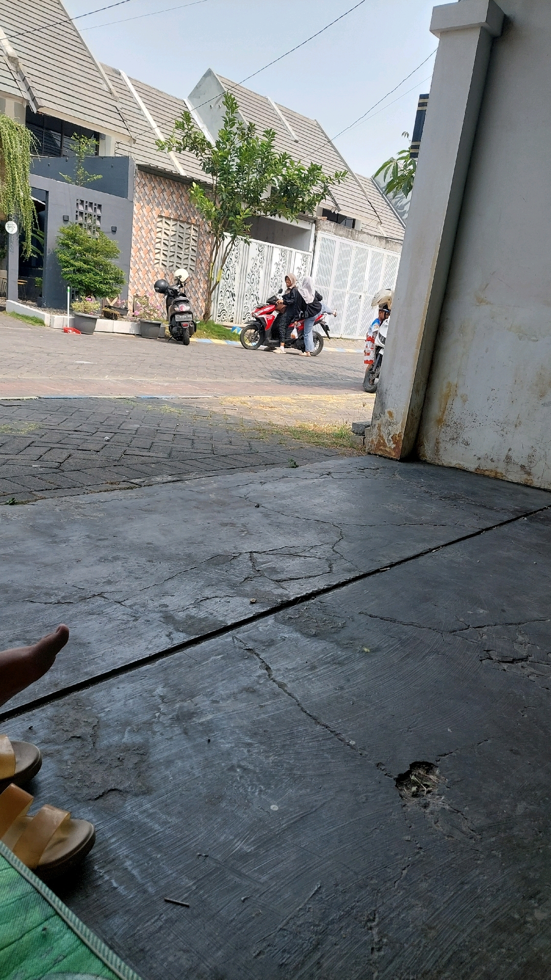 In Jimbaran Kulon, Indonesia, two young women are on a red scooter on a street. One woman wears a black hijab and a black shirt, and the other wears a white hijab and a dark blue shirt. They are stopped at a red light next to a black scooter, and a third person on a white scooter is behind them. The street is paved with cobblestones and bordered by a sidewalk. Houses with pitched roofs and white decorative fences are visible in the background, as well as a large, weathered pillar on the right side of the frame. In the foreground, a foot wearing a sandal is visible on a cracked concrete surface.