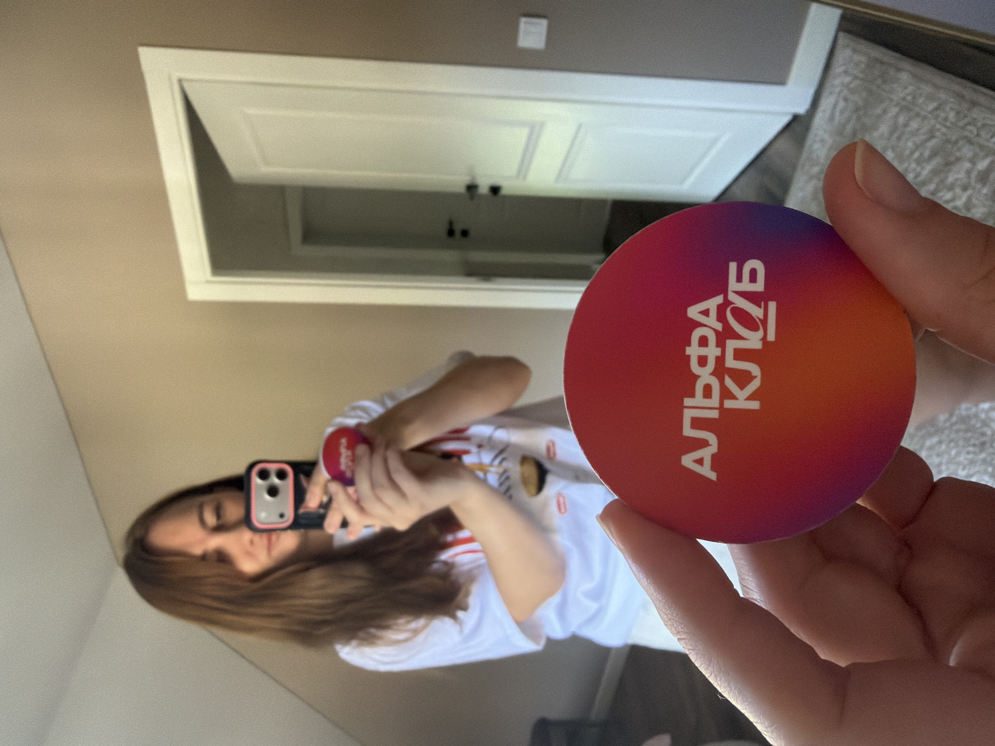 A woman is taking a selfie in a mirror, holding a phone in her left hand and a round red and purple sticker in her right hand. The sticker has white Cyrillic text that reads "АЛЬФА КЛУБ" (Alpha Club). She is wearing a white t-shirt with a graphic print and white shorts. The background shows a doorway with a partially open white door, a wall painted tan, and a rug on the floor. There is no indication of the city of Malinovka, Russia in the image.
