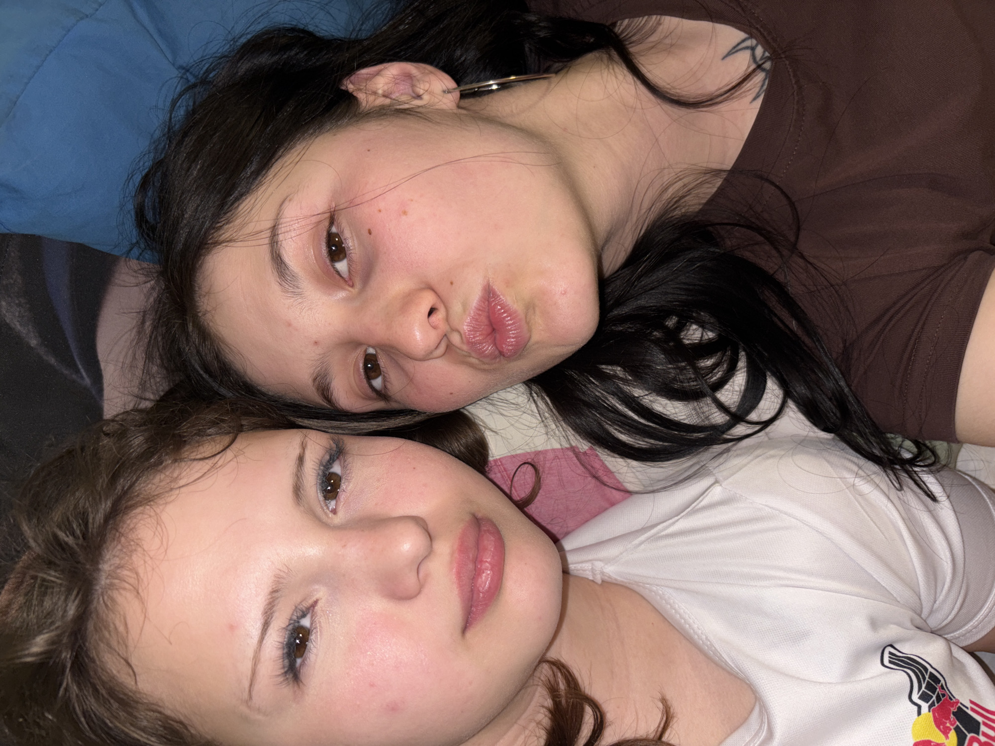 Two young women are lying down next to each other, posing for a photograph. The woman at the top of the frame has dark hair, brown eyes, and is wearing a brown shirt. She is pouting her lips slightly. The woman at the bottom has lighter brown hair and brown eyes, wearing a white shirt with a logo on the right side. The background includes a blue object and what appears to be a pink and white patterned surface behind the women. The location, Фоки, Russia, cannot be determined from the image content.