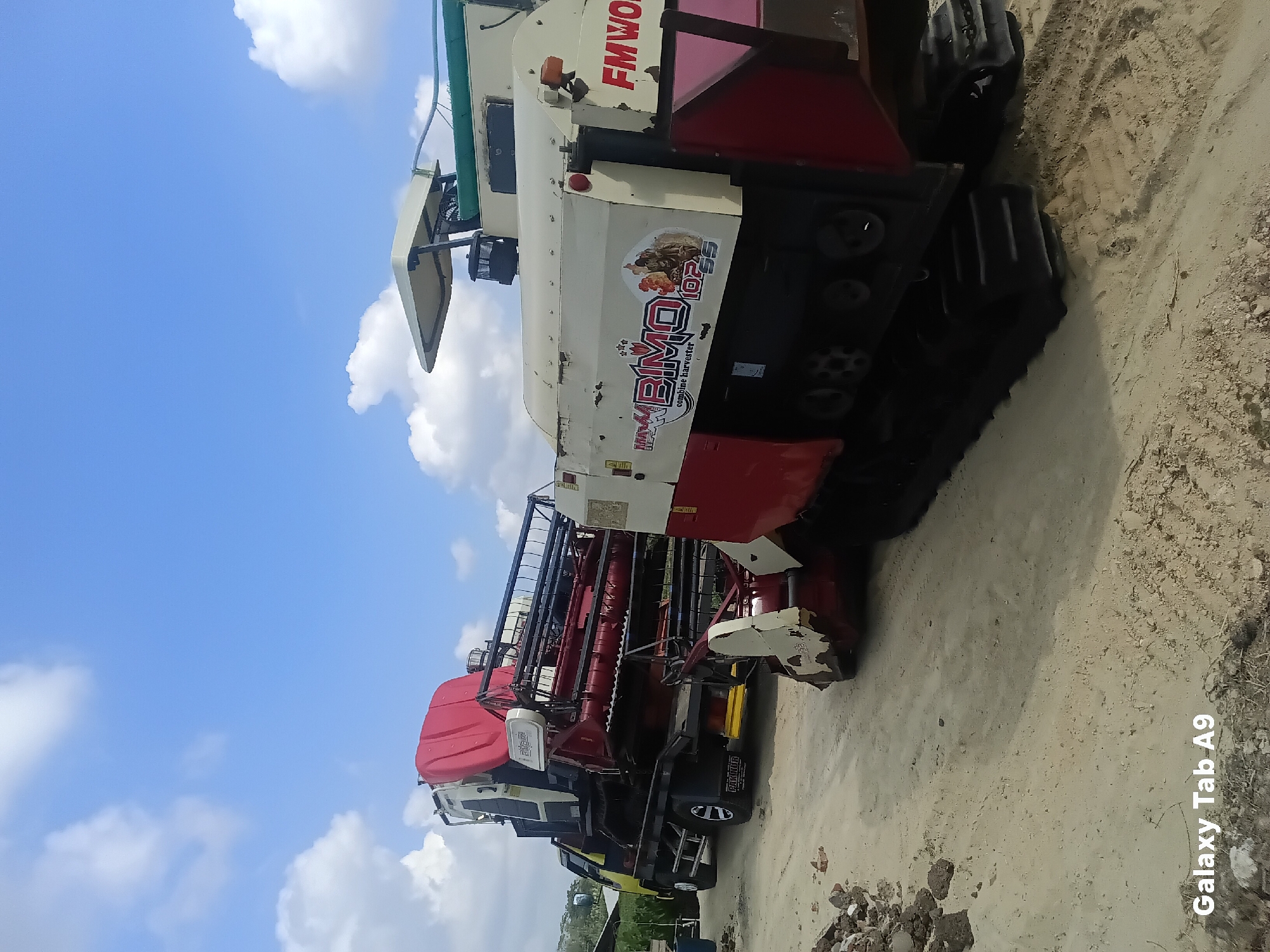 Two combine harvesters are parked on a sandy ground in Bendorejo, Indonesia. The harvester on the left is loaded onto a yellow truck. It is red and white with a red tarp on top. The harvester on the right is cream and red with a green top. The word "BIMO" is visible on the side of the harvester on the right. The sky is blue with white clouds. There are piles of debris on the ground to the left of the harvesters. The text "Galaxy Tab A9" is in the bottom left corner of the image.
